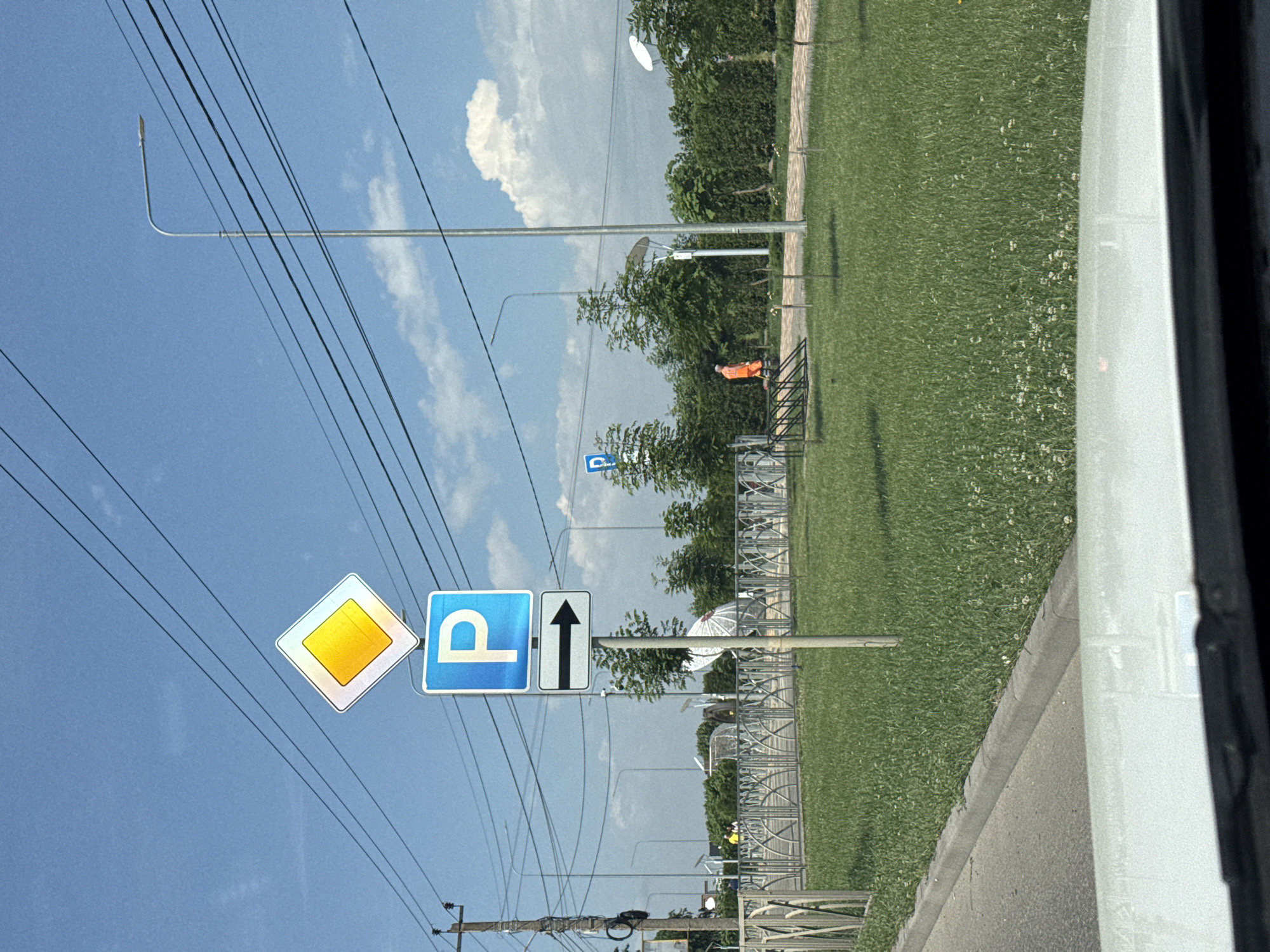 The image shows a street scene in Mikhaylovsk, Russia, on a bright, sunny day with a blue sky and scattered clouds. In the foreground, a white car's hood is visible. A paved road with a curb runs along the left side of the image, bordered by a well-maintained green lawn.
Prominently featured on the right side of the road is a traffic signpost. The top sign is a yellow diamond with a white border, indicating a main road. Below it is a blue square sign with a white letter "P" signifying parking. Underneath the parking sign is a white rectangular sign with a black arrow pointing to the right, indicating the direction of the parking area.
Further back, a paved walkway is visible, lined with trees and bushes. A person wearing an orange uniform and a hat is walking on this walkway, pushing a broom. In the background, there are more trees, fences, and streetlights, as well as some buildings and possibly a satellite dish. A metal bicycle rack is also visible on the left side of the walkway. The overall environment suggests a public area, possibly a park or a street near a commercial or residential zone.