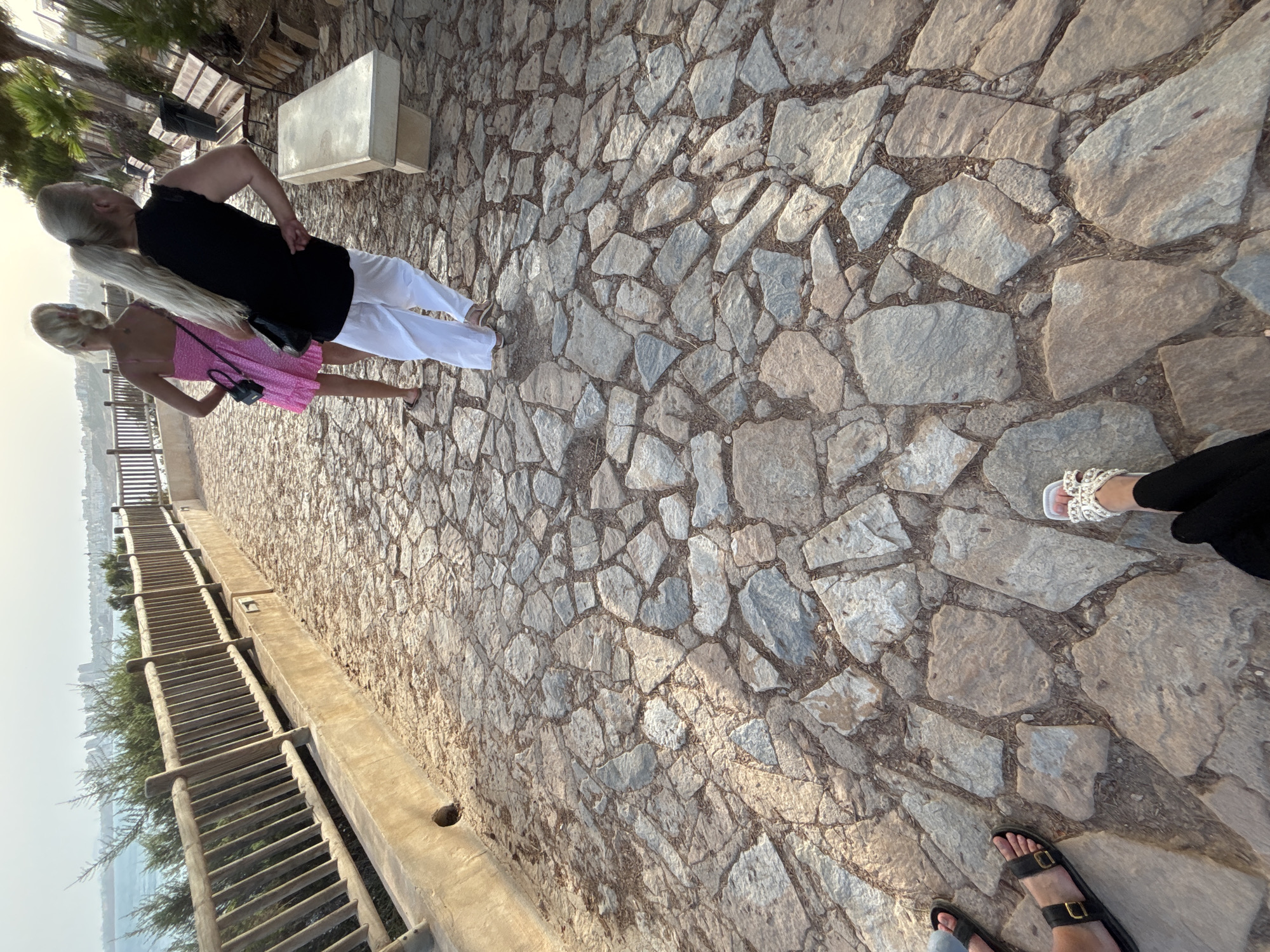 The image captures a paved walkway in Orihuela, Spain, composed of irregularly shaped stones. A wooden railing runs along the left side of the path, overlooking a body of water and a distant cityscape. Several people are visible on the walkway. In the foreground, a person's feet are clad in white sandals and black clothing. Further up the path, two women are walking away from the camera. One wears a pink dress and a black purse, while the other is dressed in a black top and white pants, also carrying a purse. Wooden benches are situated to the right of the walkway. The lighting suggests it is late afternoon or early evening.