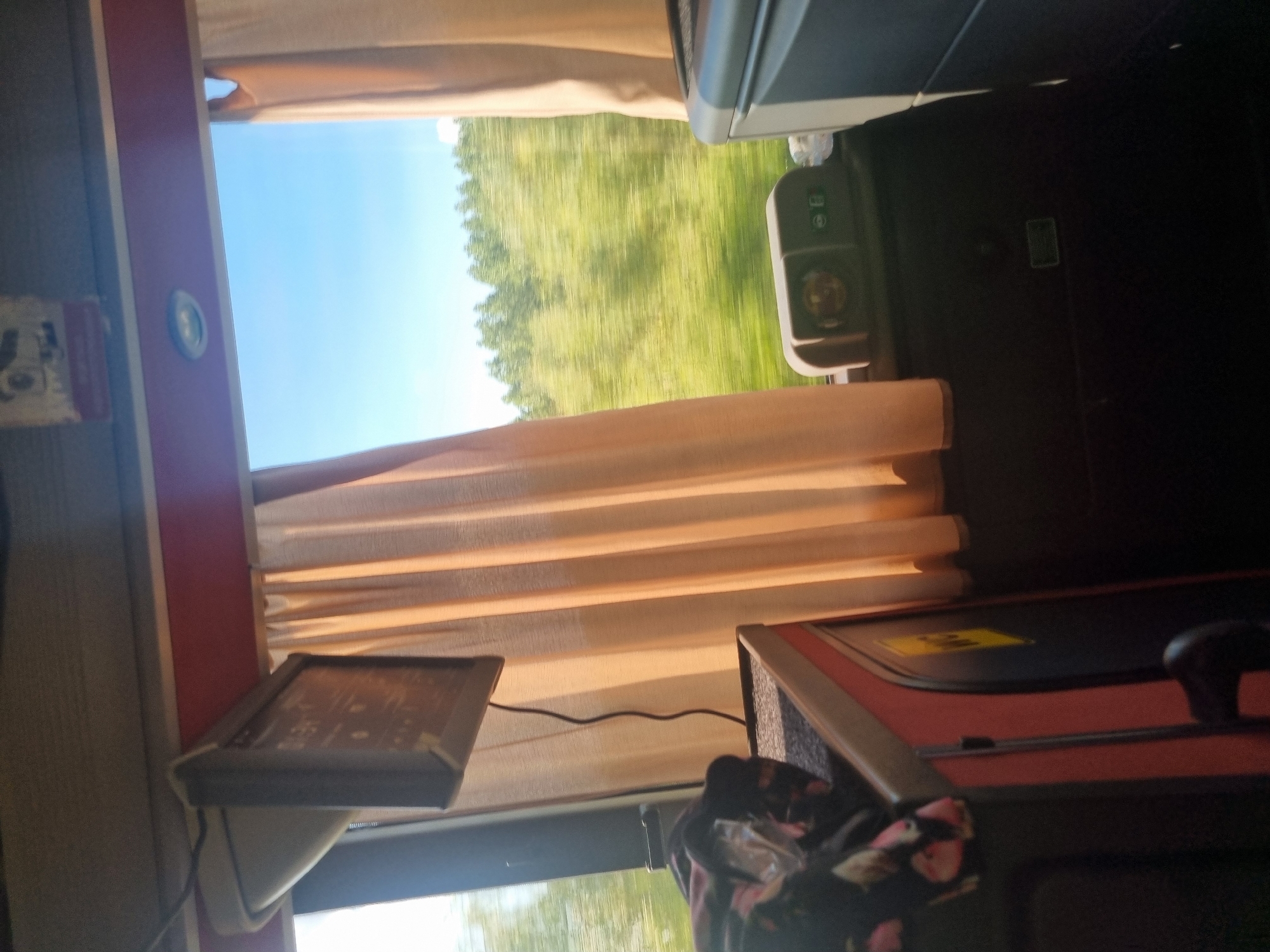 The image depicts the interior of a bus in Fényeslitke, Hungary. Sunlight streams through the window, illuminating the scene. To the left, a small television screen is mounted on the ceiling. Adjacent to it, beige curtains hang, partially obscuring the view outside. Outside the window, a blurred landscape of green trees under a clear blue sky is visible, indicating movement. In the foreground, on the right, a dark grey seatback is present. To the left, a black surface with red trim is visible, upon which a bundle of dark fabric with pink patterns is placed. The bus appears to be on a journey through the Hungarian countryside.