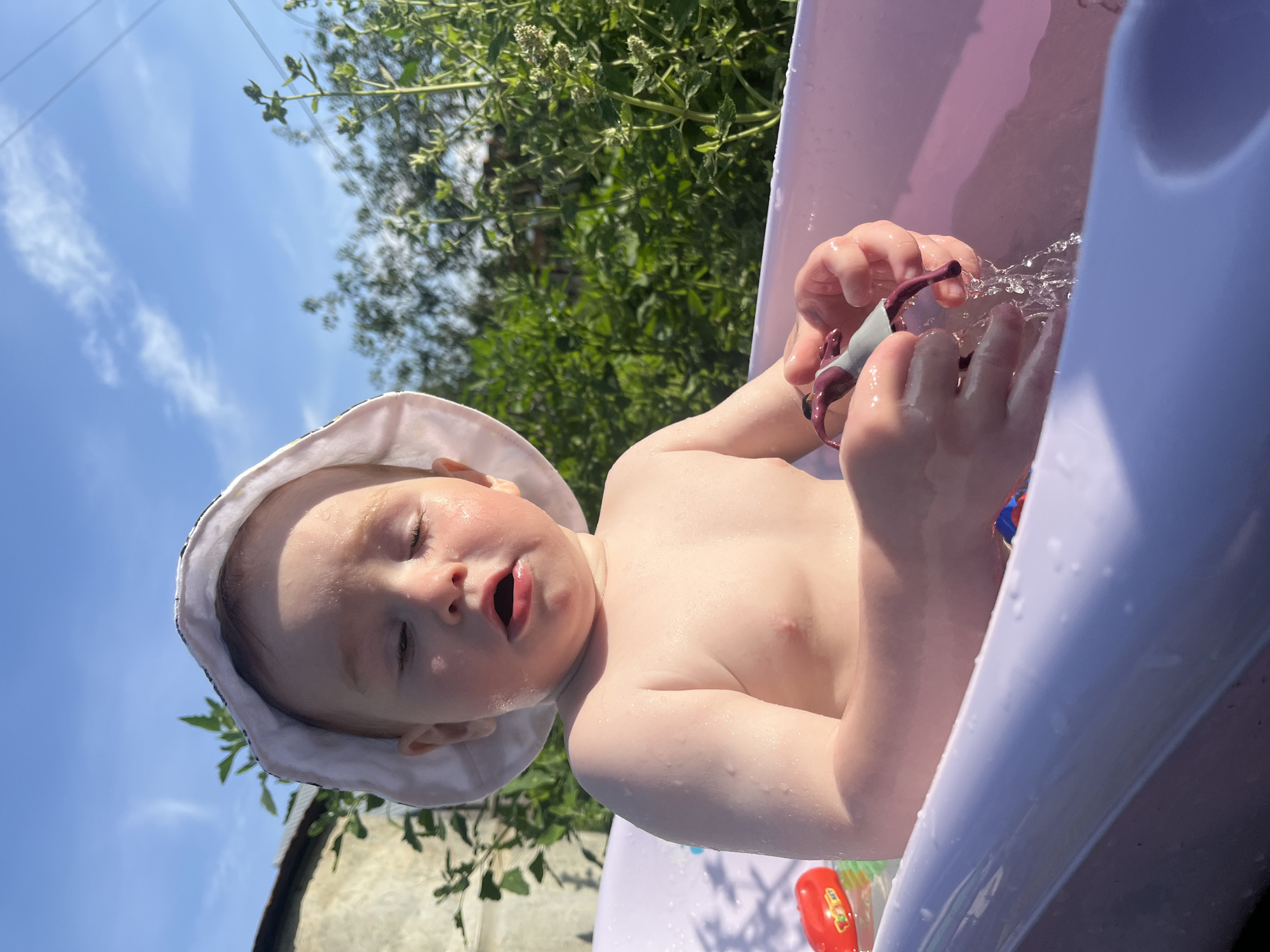 An outdoor, close-up, eye-level, medium shot captures a toddler sitting in a light purple baby bathtub filled with water. The toddler, wearing a white sun hat, has their eyes closed and mouth open. They are holding a small, dark purple and gray toy in their hands, with water splashing from it. Several other toys, including a red one with yellow and green accents, are visible in the bathtub. The background shows lush green foliage and a bright blue sky with scattered clouds. The setting appears to be a backyard in Chaikovsky, Russia.