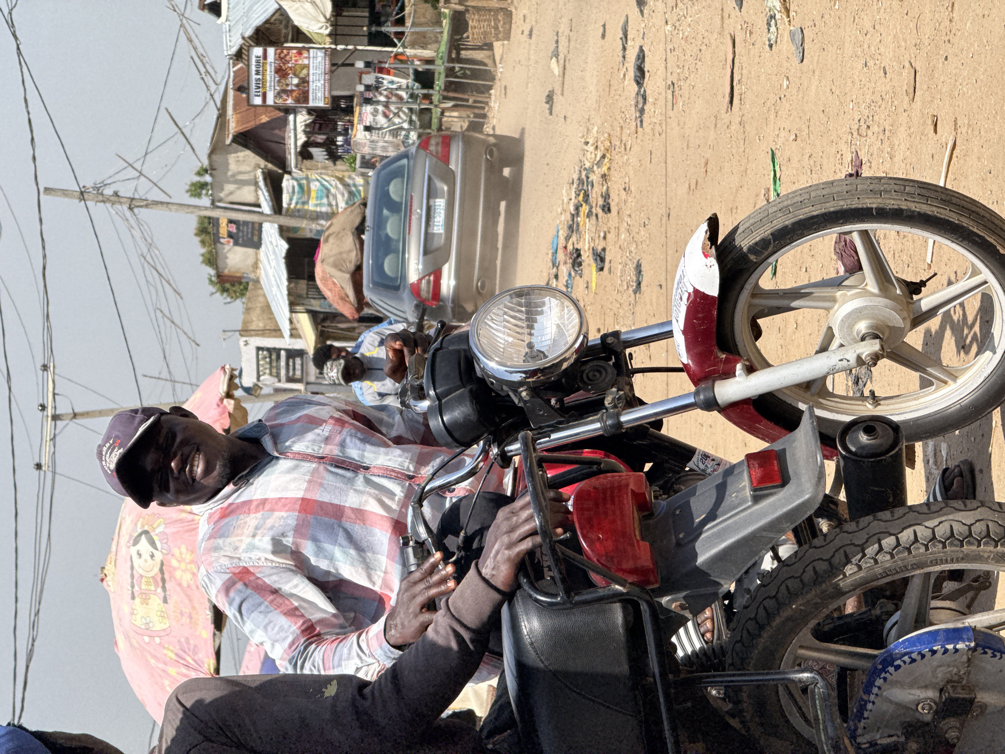The image shows a man in a red, white, and blue plaid shirt and a dark baseball cap smiling while sitting on a motorcycle. He appears to be in an outdoor market or busy street scene. In the background, there are buildings with corrugated metal roofs, stalls selling goods, and power lines. A silver car is visible driving on a dusty road, and there is trash scattered on the ground. The lighting suggests it is daytime, likely sunny. The man's expression conveys a sense of happiness. A pink umbrella with a cartoon drawing of a girl on it is visible behind him. Text on a sign in the background reads "ELVIS MORE (Supreme) Family".