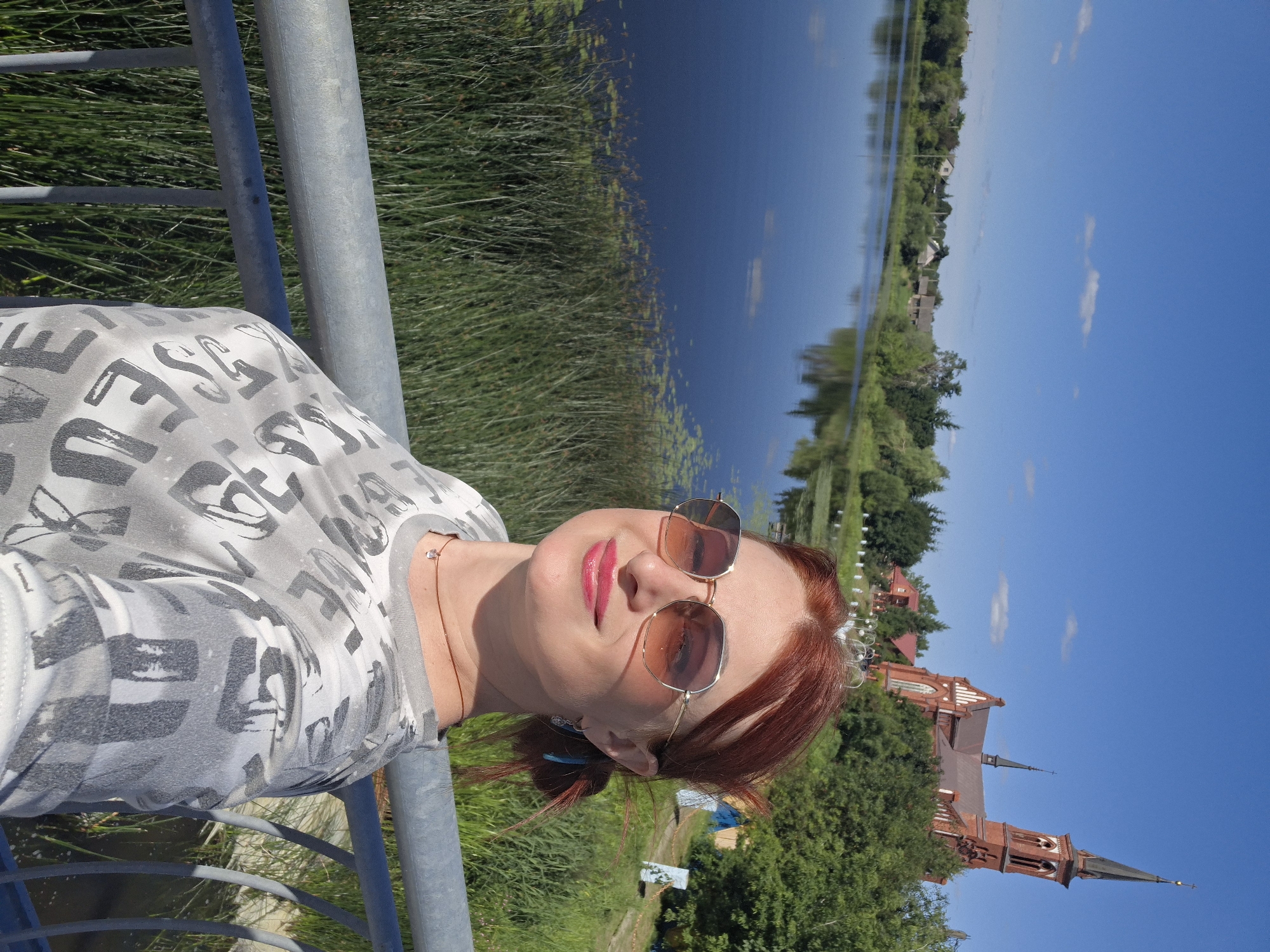 The image shows a woman with red hair, wearing sunglasses and a grey t-shirt with a patterned design, standing on a bridge or railing. She is smiling and looking at the camera. In the background, there is a body of water with reeds and lily pads. Across the water, there are trees, houses, and a prominent red brick church with a tall spire. The sky is blue with a few scattered clouds. This scene is likely in the city of Pastavy, Belarus, given the presence of the church and the rural/waterfront setting. The overall atmosphere is bright and sunny, suggesting a pleasant day.