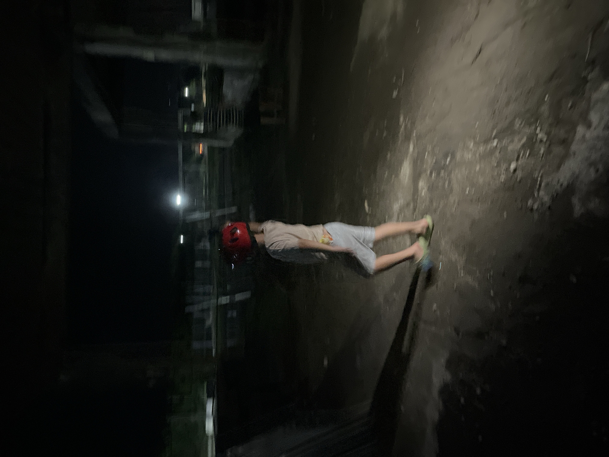 A child wearing a red helmet walks in an outdoor, dimly lit area at night. The child is wearing a light-colored t-shirt, grey shorts, and green flip-flops. The ground is uneven and appears to be dirt or gravel. In the background, there are structures that suggest an industrial or abandoned site, with concrete pillars and some fencing visible. The overall atmosphere is dark and somewhat desolate, illuminated by sparse artificial lighting from a distance. The location is Poti, Georgia.