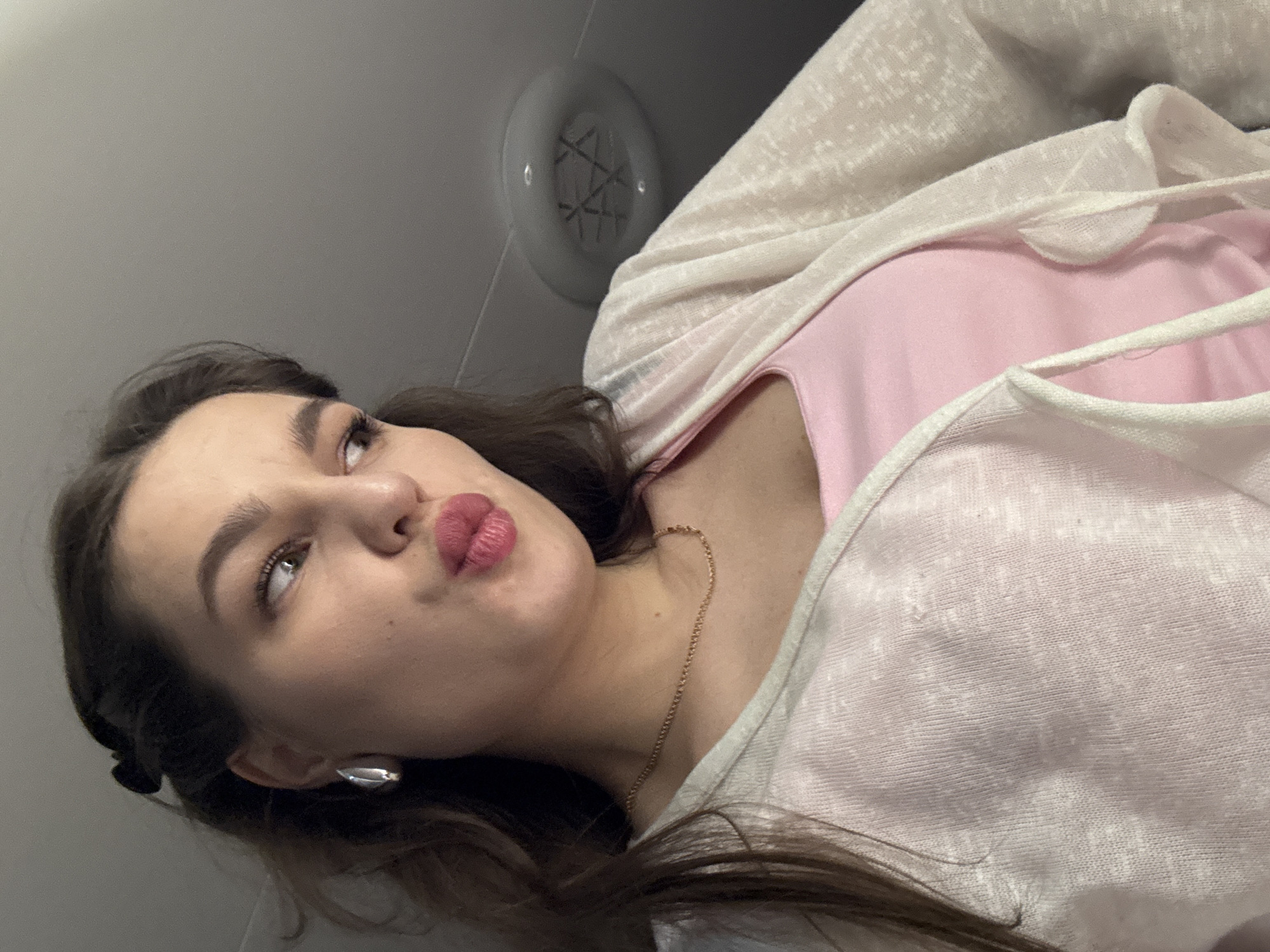 A woman is photographed from a low angle, looking upwards and to the left. She has brown hair, fair skin, and is wearing an earring on her left ear. She is wearing a pink top and a light-colored cardigan. A thin, gold necklace is visible around her neck. The background consists of a light gray ceiling with a round, white vent. It is not possible to determine from the image if this scene is in Chaikovsky, Russia, or anywhere else.