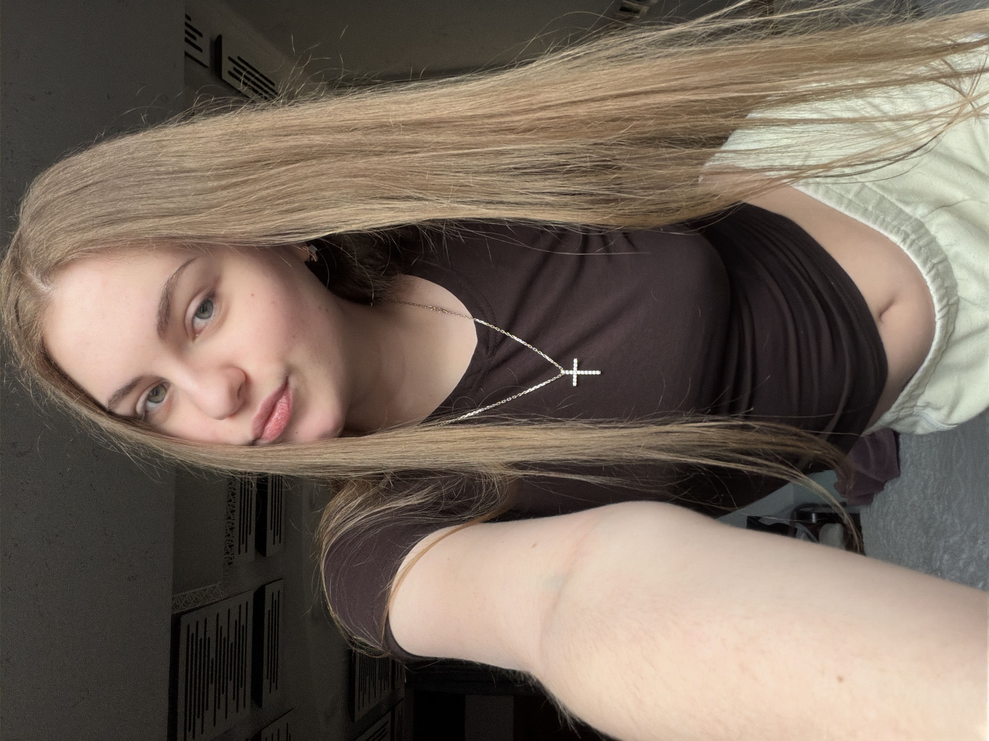 This is a selfie of a young woman with long, blonde hair. She is wearing a dark brown t-shirt and light green sweatpants. A silver necklace with a cross pendant is visible around her neck. Her left arm is extended towards the camera, taking the picture. The background appears to be an indoor setting with textured ceilings and a wall with ventilation grilles. The lighting is dim, creating shadows in the background. There is no indication of the city of Dnipro, Ukraine, in the image itself.