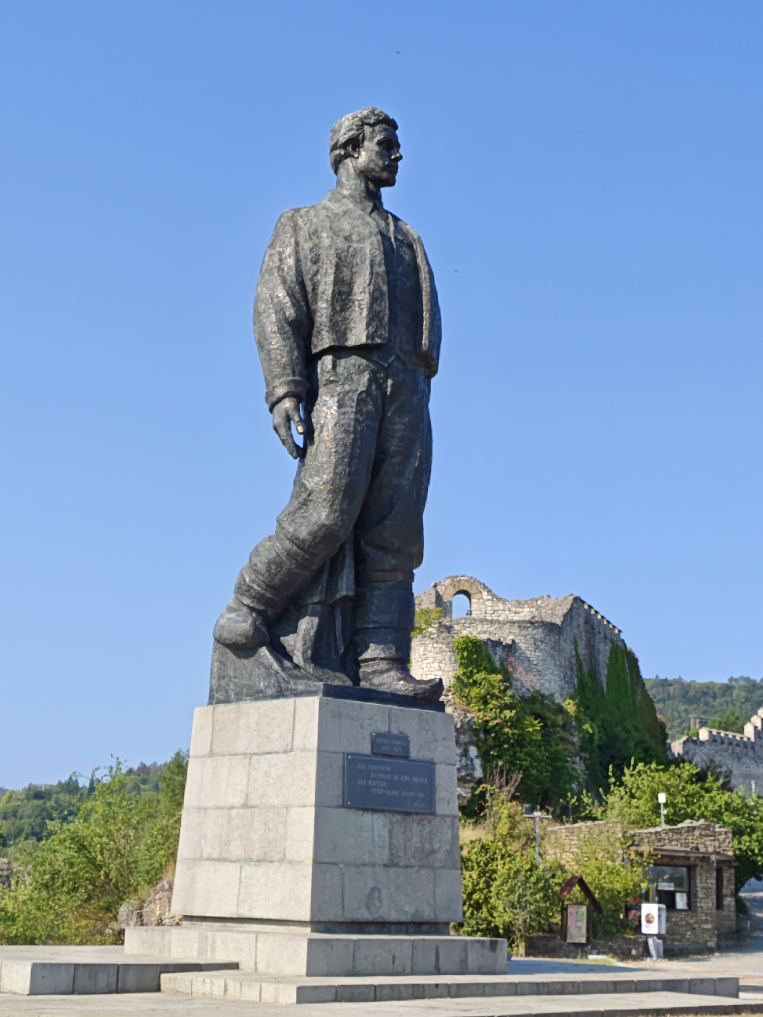The image shows a large bronze statue of Vasil Levski, a Bulgarian national hero, standing on a pedestal in Lovech, Bulgaria. The statue depicts Levski in a standing pose, wearing a traditional Bulgarian coat and trousers. The pedestal is made of stone blocks. In the background, the ruins of the Lovech Fortress, also known as Hisarya Fortress, are visible. The fortress is covered in greenery, and parts of its stone walls and arched windows are discernible. The sky is clear and blue. The overall scene is one of historical significance and natural beauty.