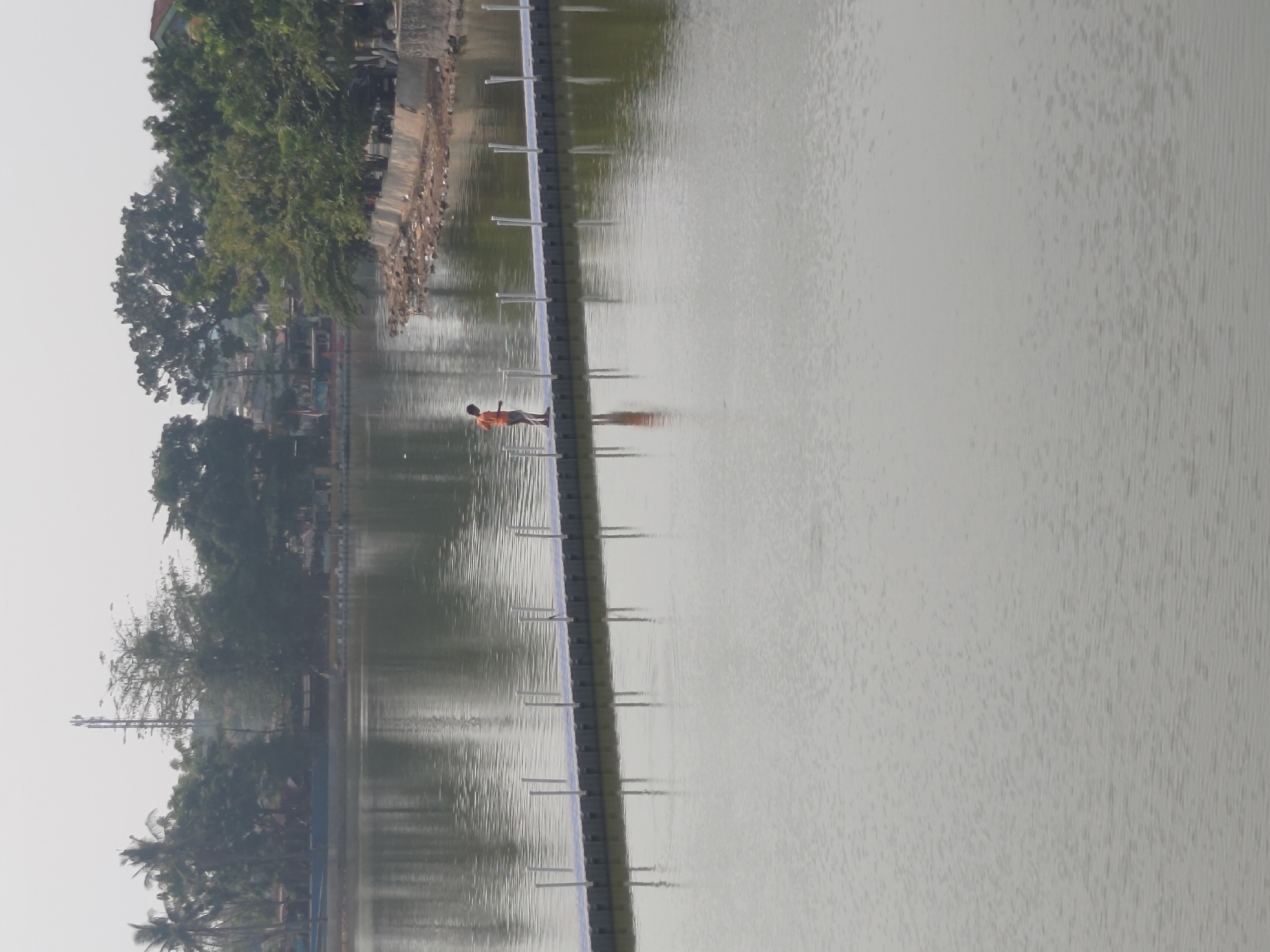 A boy in an orange shirt stands on a floating walkway extending into a calm body of water, likely a lake or reservoir, in Depok, Indonesia. The walkway is made of modular plastic blocks and has white posts lining its sides. In the background, lush green trees line the shore, with some buildings and a cell tower visible beyond them. The sky is overcast, casting a muted light on the scene. The water reflects the surrounding trees and walkway.