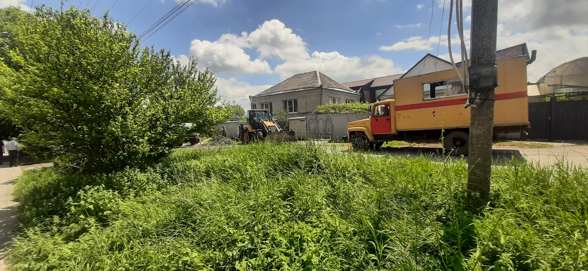 This image depicts an outdoor scene in Yessentukskaya, Russia, featuring a residential area with construction or maintenance work underway. In the foreground, dense, tall green grass and foliage dominate, with a paved path visible to the left. Beyond the grass, a yellow truck with a red door and a red stripe is parked, its driver visible in the cab. To the left of the truck, an orange backhoe loader is positioned near a light-colored brick building, suggesting ongoing construction or excavation. Several other buildings, including a residential structure with a tiled roof, are visible in the background, along with utility poles and overhead wires. The sky is partly cloudy with bright sunlight.