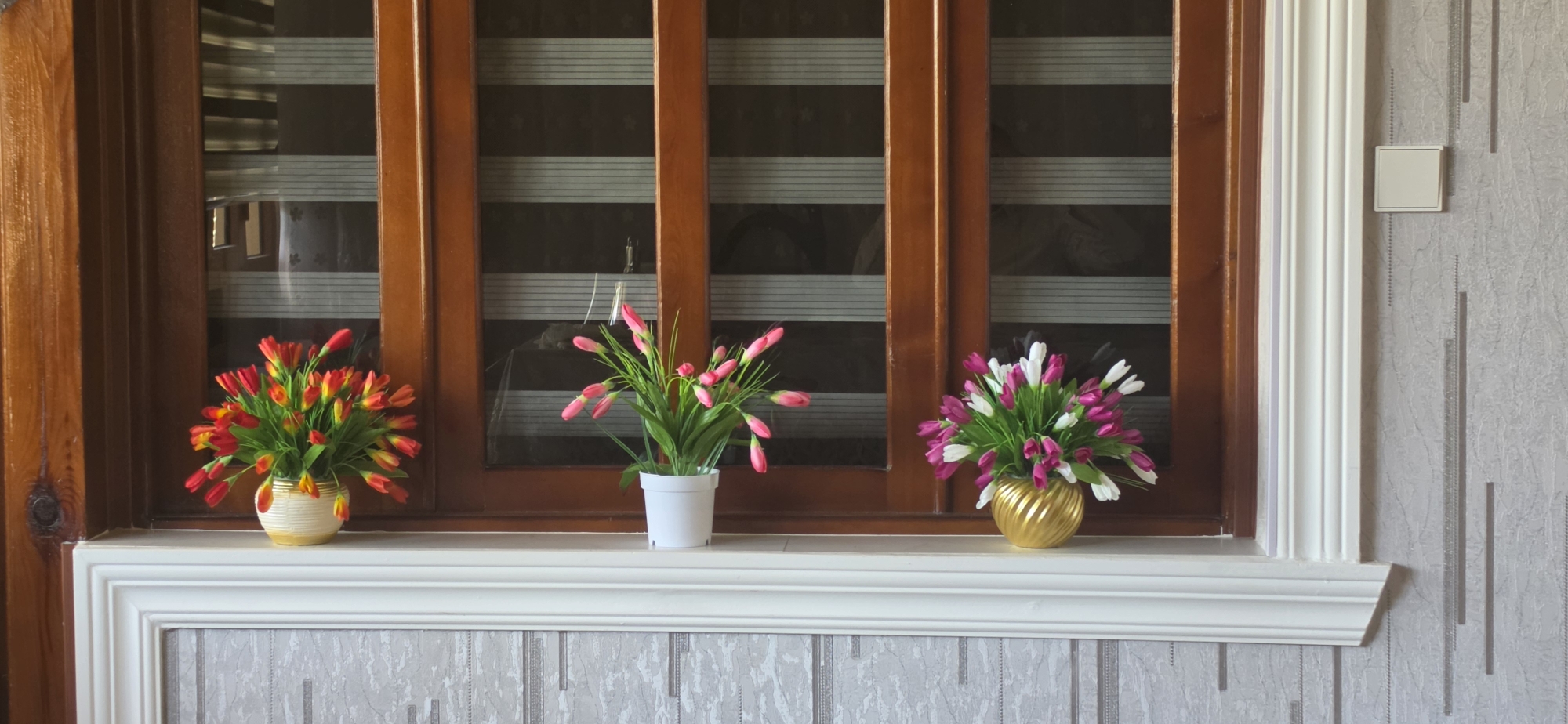 In Uychi, Uzbekistan, a windowsill displays three artificial flower arrangements in front of a wooden-framed window with horizontal blinds. The leftmost arrangement features red and yellow tulips in a white and tan patterned vase. The central arrangement consists of pink tulips in a white pot. The rightmost arrangement shows pink and white tulips in a shiny gold vase. The windowsill is topped with white molding, and the wall to the right is covered in grey textured wallpaper with vertical decorative lines. A white square electrical outlet is visible on the wall.