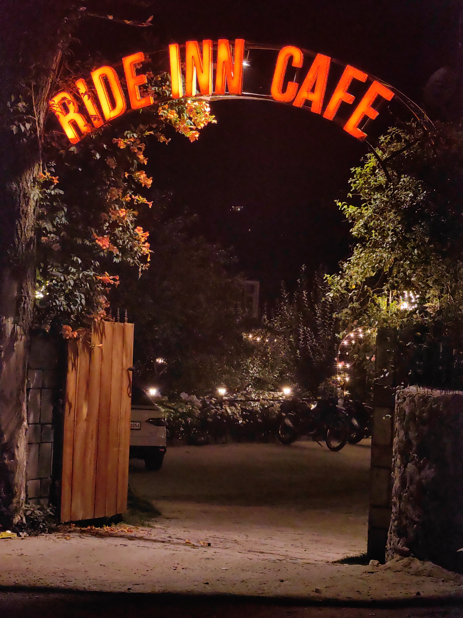 An outdoor nighttime scene at "RIDE INN CAFE" in Barheta, India. A bright red neon sign spelling "RIDE INN CAFE" is arched over an entrance. Orange flowers and greenery drape around the sign and entrance. To the left, a rustic wooden gate is ajar, revealing a white car parked behind it. To the right, stone pillars frame the entrance. Beyond the entrance, several motorcycles are parked, and string lights illuminate the foliage and pathway. The ground is paved with gravel. The overall atmosphere is warm and inviting, with the glowing sign and ambient lighting creating a welcoming ambiance.