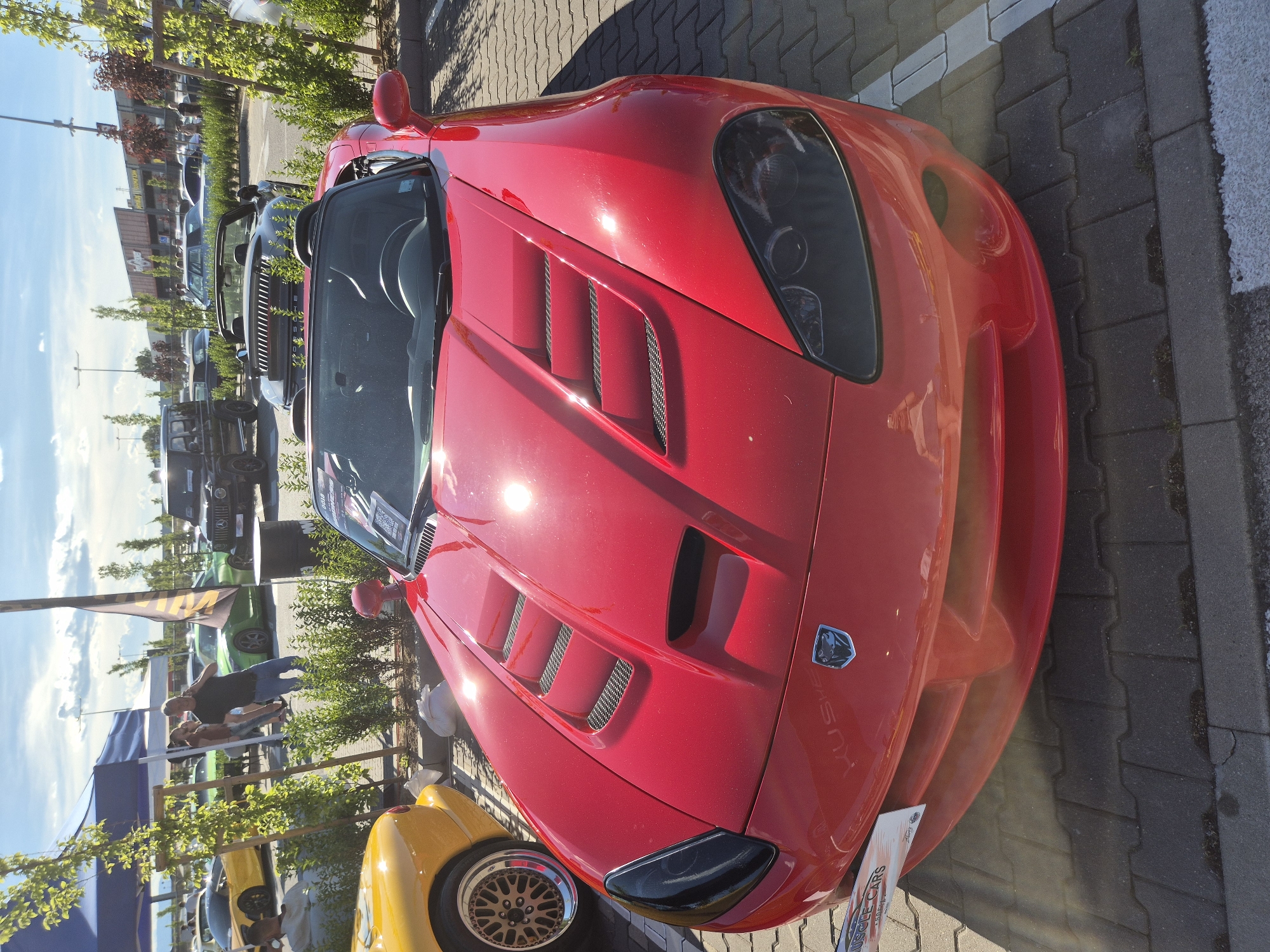 A red convertible Dodge Viper is parked in a lot with other vehicles, possibly at a car show. The car is in the foreground, facing left, with its hood prominently displayed. The hood has several air vents and a distinctive shape. The car has black headlights. In the background, a yellow sports car is parked to the left of the Viper. Further back, several other cars, including a black SUV, are visible. People are scattered throughout the background, some walking and others standing near the cars. The scene appears to be outdoors during the day, with a bright sky and sunlight casting shadows. The ground is paved with interlocking bricks. A blue tent is visible on the left side of the image.