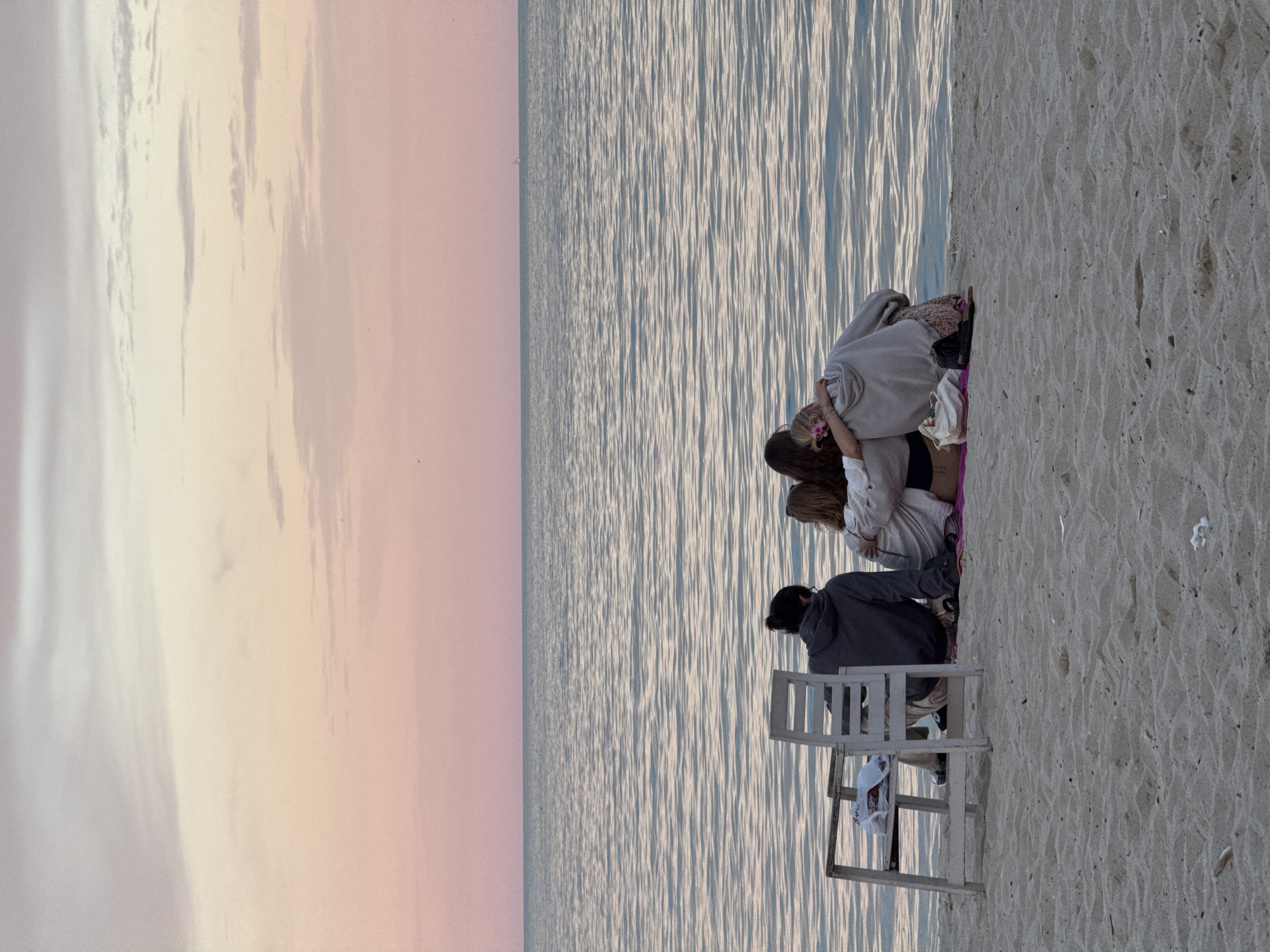 Four individuals are seated on a sandy beach, facing the sea. The sky above is painted with soft hues of pink and pale orange, indicative of either sunrise or sunset. The sea itself is calm, with gentle ripples reflecting the colors of the sky.
To the left, a white wooden chair is positioned on the sand. A person wearing a grey hooded sweatshirt and dark pants is seated on the beach, slightly to the right of the chair. This individual is facing away from the camera, towards the sea.
Clustered together in the center of the image, the other three individuals are also seated on the beach, facing the water. They are close together, with arms around each other, suggesting companionship. Two of them are wearing hoodies, and one is wearing a patterned skirt. A pink blanket is spread on the sand beneath them, and a white handbag and dark shoes are placed nearby.
The location is identified as the Varna Black Sea coast in Varna, Bulgaria.