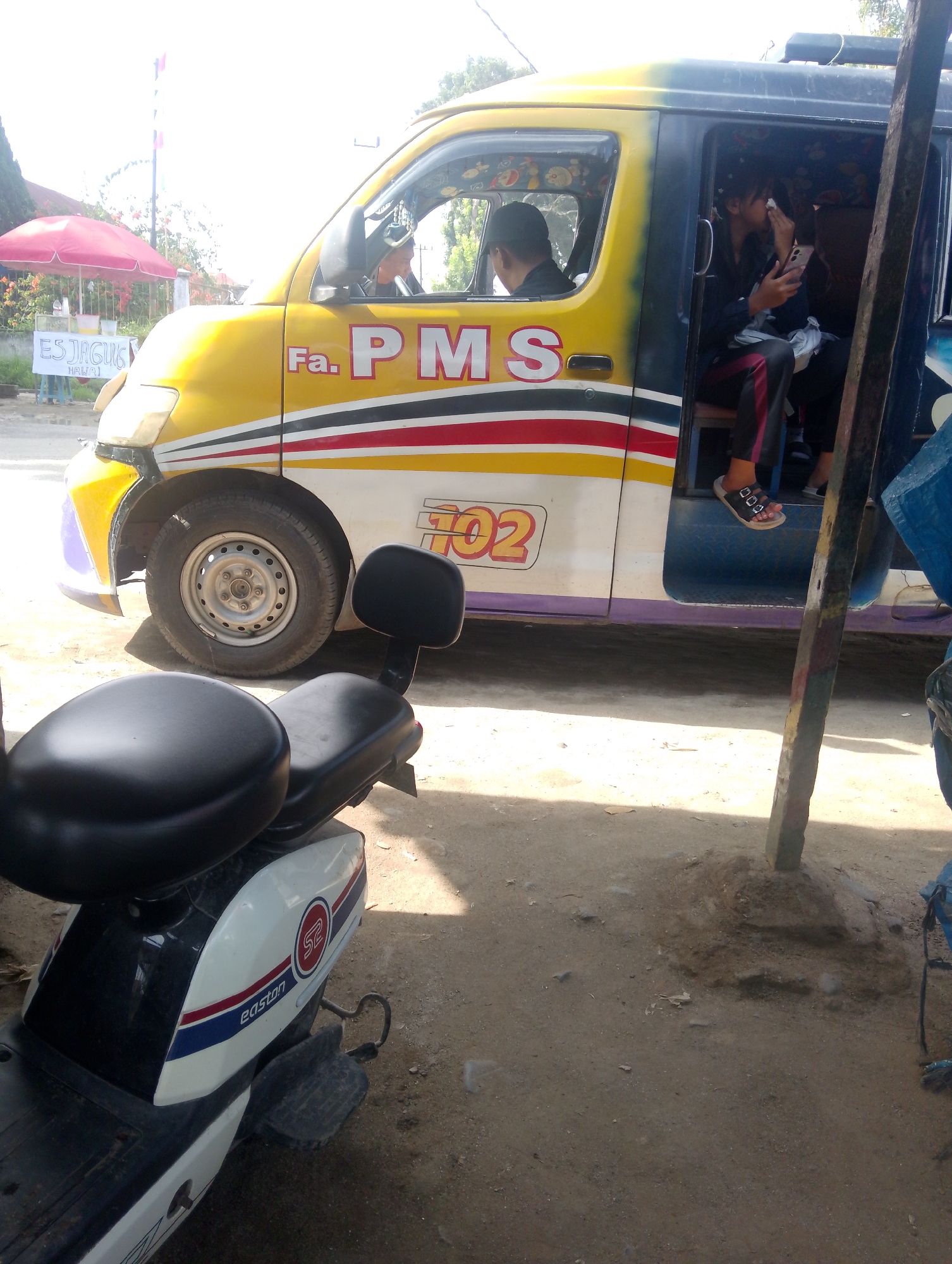 In Serbelawan, Indonesia, a yellow and white vehicle, possibly a van or a modified car, is parked. It has the words "Fa. PMS" and "702" painted on its side. A person is visible driving the vehicle, and another person is seated inside. In the foreground, a white and black scooter is parked. The scene appears to be outdoors, with a dusty or unpaved ground. There is also a red umbrella and a sign that says "ES JAGUNG" in the background.
