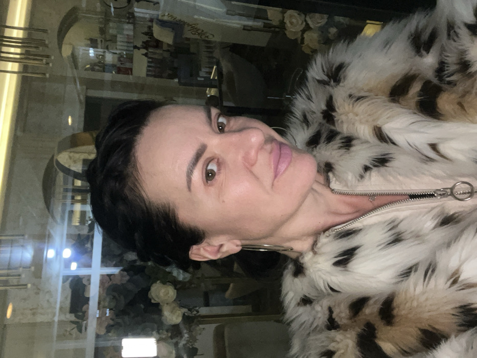 The image displays a head-and-shoulders selfie of a woman, aged approximately 40-50 years, situated indoors within the Smederevska Palanka Municipality, Serbia. The woman has dark hair styled away from her face, prominent brown eyes, and light skin with visible makeup, including pink lipstick. She is adorned with large silver hoop earrings. Her attire consists of a faux fur coat, predominantly white and cream with distinct mottled dark brown or black patches. Beneath the fur, a light pink or beige garment with a silver zipper and a circular pull tab is visible. The background, observed through reflection in a glass or mirrored surface, reveals an interior commercial space, likely a salon or beauty establishment. Visible reflections include shelves stocked with various product bottles, a partially legible sign featuring stylized text, an arrangement of light-colored artificial flowers, and modern interior lighting, including a vertical illuminated panel and a metallic chandelier-like fixture.