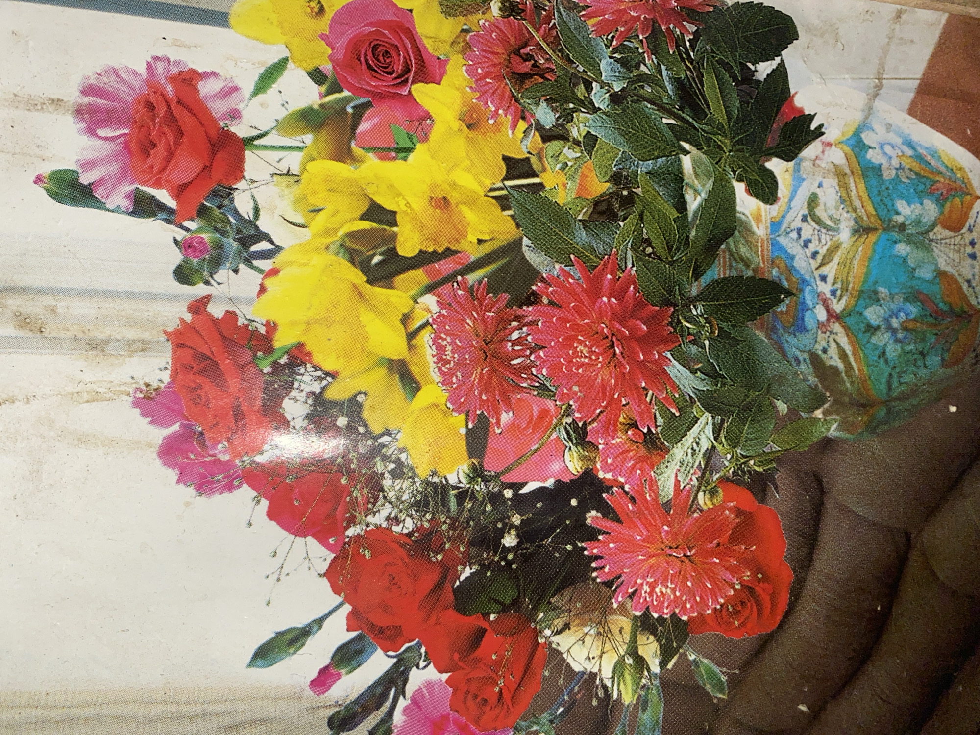 This media file is a close-up, overhead shot of a vibrant bouquet of flowers, possibly placed in a patterned vase. The bouquet features a mix of red roses, yellow daffodils, and bright red dahlia-like flowers, interspersed with lush green leaves and delicate baby's breath. The overall impression is one of abundance and natural beauty.
The flowers are arranged in a dense cluster, with some petals overlapping and others reaching out from the bouquet. The colors are saturated and bold, suggesting a sunny environment or high-quality printing. The background is a light, textured surface that appears to be a wall or tablecloth, with faint horizontal lines suggesting wood grain or a woven fabric. To the right, a portion of a hand is visible, with dark skin and a curved form, suggesting the bouquet is being held or presented. This hand is partially obscured by a colorful, patterned fabric, possibly part of the vase or a decorative element.
There is no visible text in the image. The lighting is bright and even, casting minimal shadows, which implies it was taken in good daylight. The setting is neutral, with no specific location cues beyond the flowers and the hand. The mood is cheerful and celebratory, evoked by the bright colors and the fullness of the floral arrangement. The scene appears to be a still life, focusing on the aesthetic appeal of the flowers.