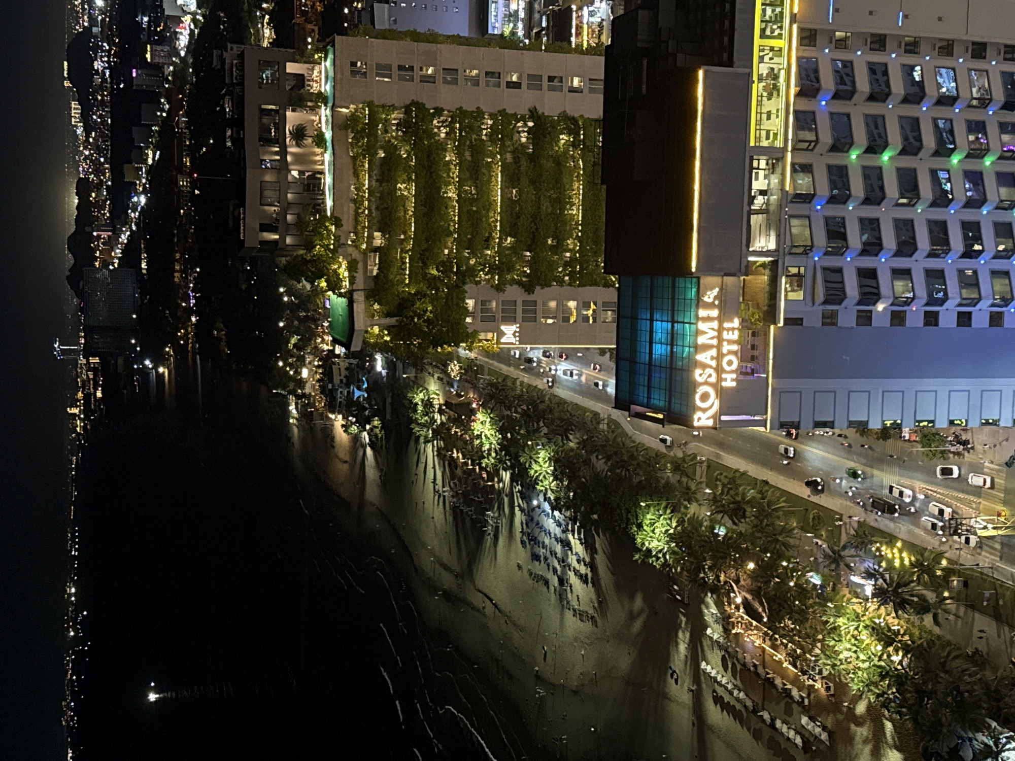 This is a nighttime aerial or high-angle view of a vibrant coastal city. The scene is dominated by artificial lights against a dark sky.
On the left, a dark body of water stretches into the distance, with a long, illuminated coastline visible across it, suggesting a bay or ocean with another part of the city or landmass beyond.
In the mid-ground, a wide sandy beach is lined with numerous palm trees. The sand appears wet, reflecting some of the bright lights from the shore. Rows of beach chairs or loungers are visible on the sand, illuminated by string lights and spotlights.
To the right of the beach runs a busy street, with several cars, both moving and parked, illuminated by streetlights and their own headlights. Further to the right, a series of multi-story buildings line the street. One prominent building features extensive vertical green gardens covering its facade, illuminated by warm yellow lights. Another large, modern hotel building, identifiable by the text "ROSAMIA HOTEL" on its facade, is brightly lit, with many windows displaying interior lights and some exterior architectural lighting in blue and green hues.
The atmosphere is lively and urban, characteristic of a bustling tourist destination at night. The numerous lights create a striking contrast against the dark surroundings. The location context indicates this is in Đà Nẵng, Vietnam.