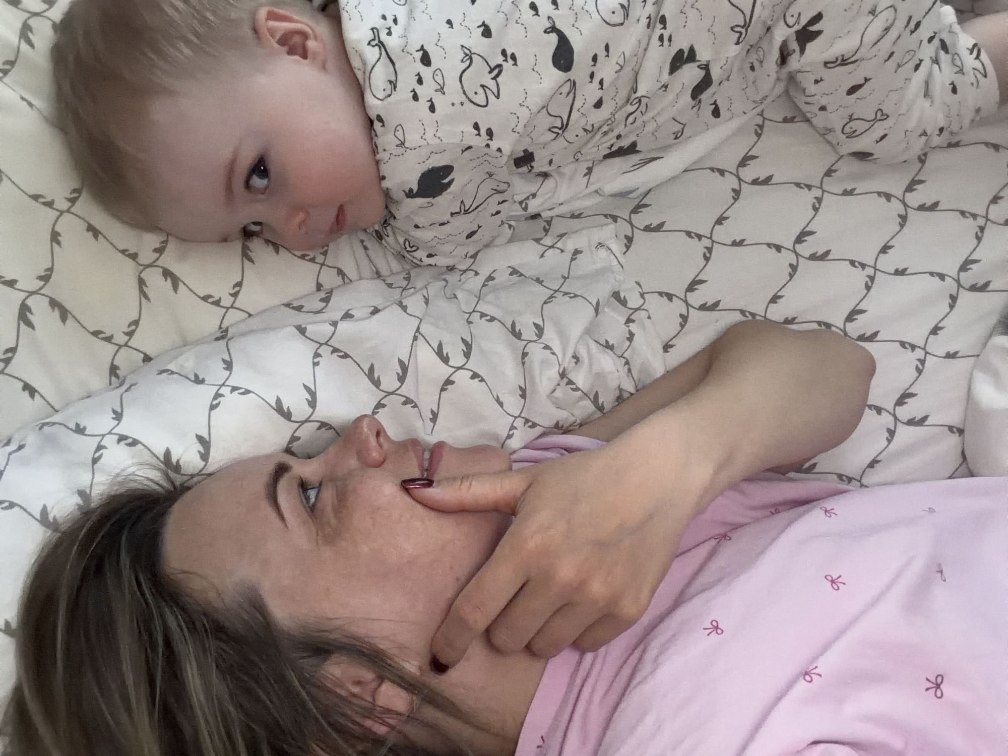 A woman and a baby are lying on a bed. The woman is lying on her back, wearing a pink top and has brown hair. She is holding her hand to her face. The baby is lying on the bed next to the woman and is wearing a white top with black drawings on it. The bed has a white sheet with a geometric pattern. The location cannot be determined without additional information.