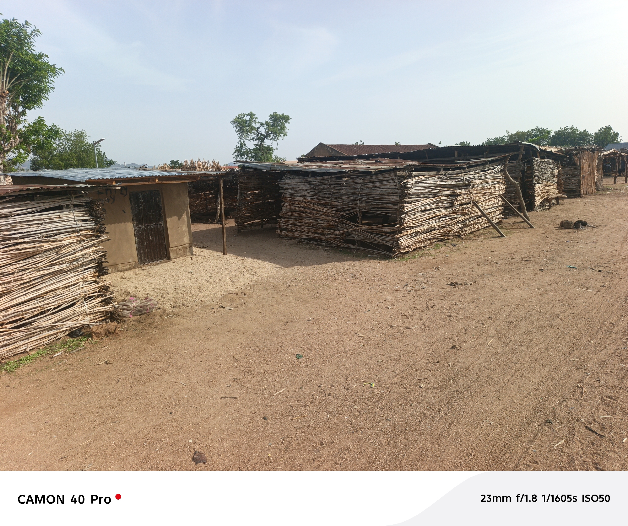 The image depicts a dirt road in Langtang, Nigeria, lined with several small, rudimentary structures. These structures appear to be made from woven branches or sticks, with some having corrugated metal roofs. One structure has a small door. The ground is reddish-brown dirt, and there are tire tracks visible in the dirt road. The sky is overcast. In the background, some trees and possibly hills are visible. There are no people visible in the immediate foreground, but a few figures can be faintly seen in the distance near the last structures. The scene appears to be a rural or underdeveloped area.