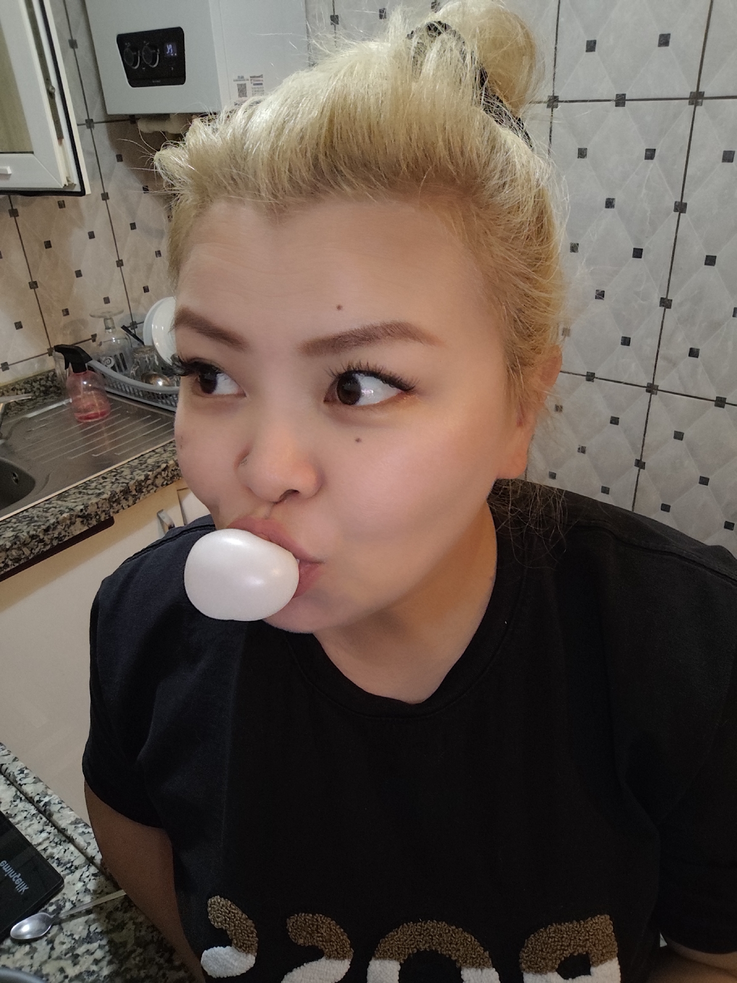 A woman with blonde hair in a bun is blowing a white bubble with gum in her mouth. She has brown eyes and is wearing a black t-shirt with white and brown patches. The background shows a tiled wall, a sink, and some kitchen appliances. The image was likely taken indoors. While the location is Sarıyer, Turkey, the photograph was taken indoors, not outdoors.