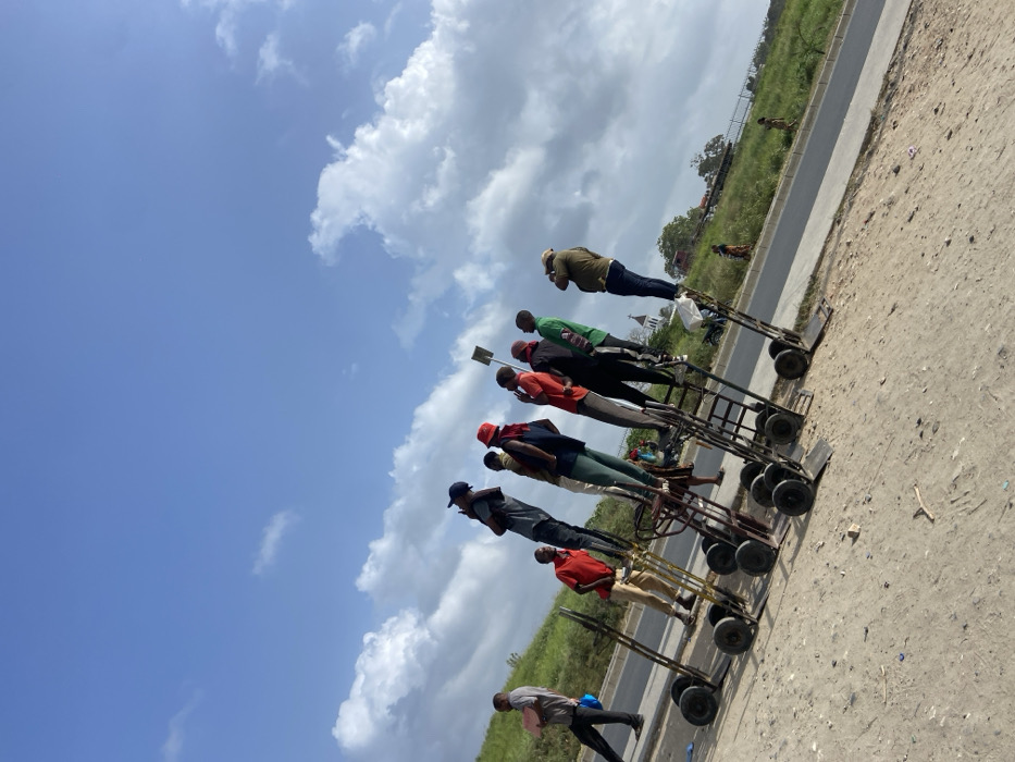 This image, taken in Dar es-Salaam, Tanzania, depicts a daytime scene under a bright, partly cloudy sky. The photograph shows a line of at least eight men standing upright beside a series of metal handcarts on a dirt and gravel roadside.
The men are casually dressed in various shirts, trousers, and some wear caps or hats. They appear to be waiting or gathered, suggesting they might be porters, vendors, or laborers who use these handcarts for transporting goods. The handcarts themselves are sturdy, multi-wheeled trolleys designed for manual transport.
To the left of the men and their carts runs a paved road or pathway. Beyond this road, there is a stretch of green grass and vegetation, with some indistinct structures and possibly a few small animals visible in the distance. In the lower left foreground, one man is captured in motion, walking towards the left side of the frame, carrying a light-colored item in his hands.
The scene conveys a sense of everyday activity in a semi-urban or roadside setting. The lighting indicates it is daytime, with scattered white and grey clouds against a blue sky. There is no visible text in the image.