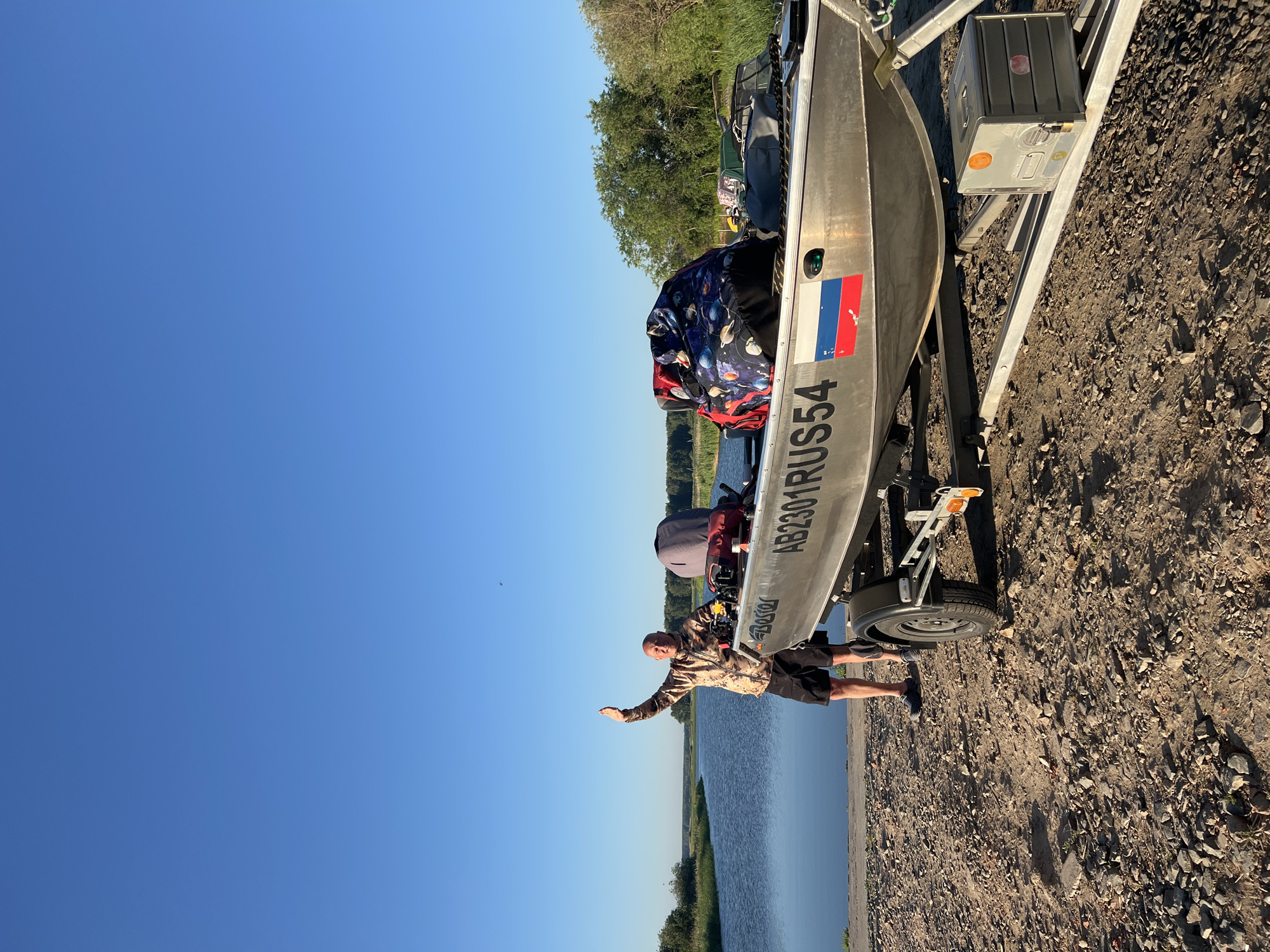 A bald man wearing a camouflage jacket and black shorts stands on a gravelly shoreline next to a boat on a trailer. He is waving his right hand, which is raised above his head. The boat, made of aluminum, has the registration number "AB2301RUS54" and a Russian flag sticker on its side. There are several bags and a grey cover inside the boat. The water of a river or lake is to the man's left, and trees and vegetation line the opposite bank. The sky is clear and blue. The scene appears to be in Novopichugovo, Russia.
