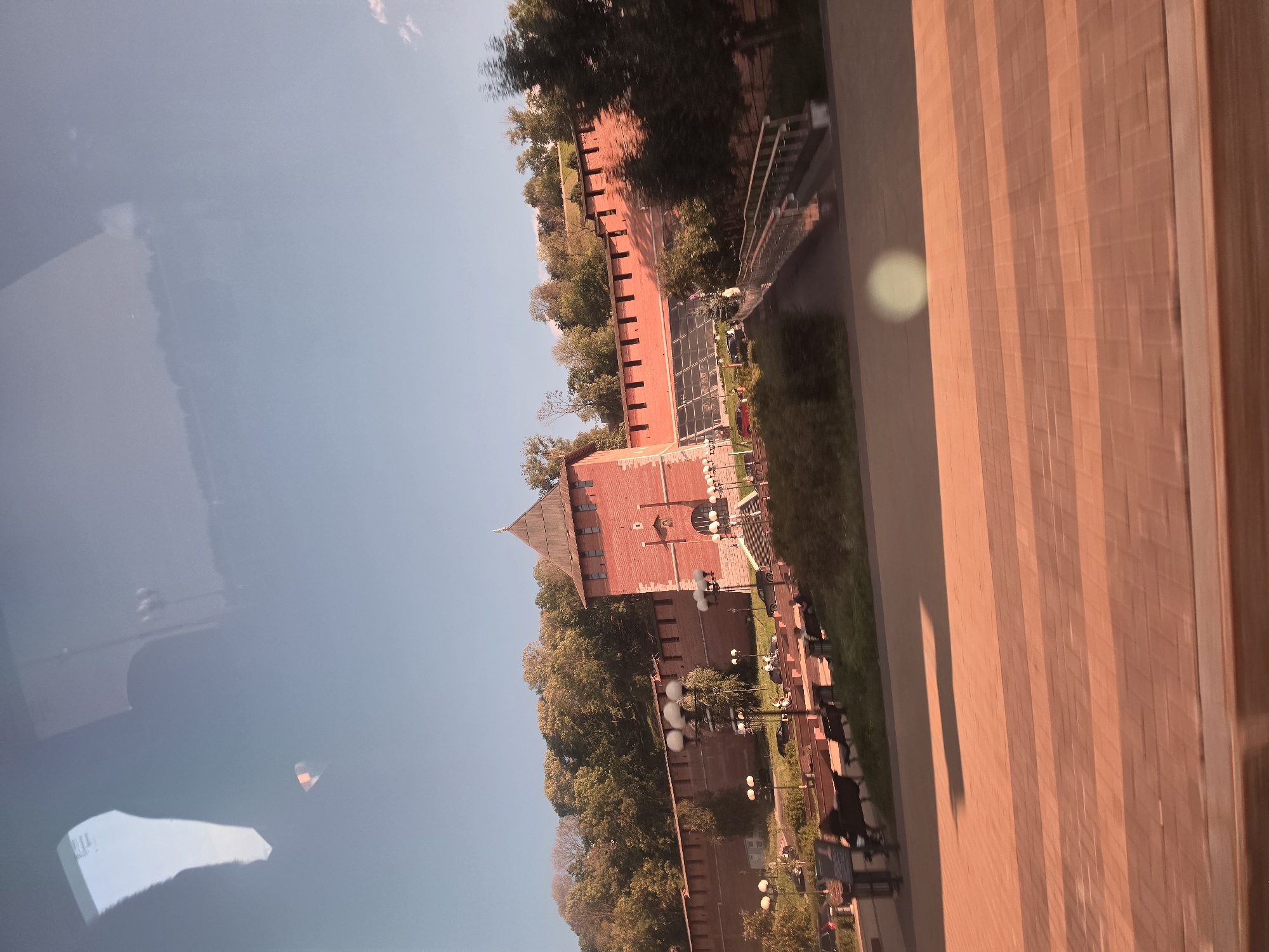 The image shows a view of the Nizhny Novgorod Kremlin, a historic fortress in Nizhny Novgorod, Russia. The Kremlin wall, constructed of red brick, stretches across the scene, topped with crenellations. A prominent red brick tower with a wooden roof stands near the center. In front of the wall, there is a pathway with lampposts and benches, some occupied by people. Beyond the path, a paved area with a brick pattern is visible in the foreground, with long shadows indicating the time of day. Trees and greenery surround the Kremlin walls. The sky above is clear and blue. The photograph appears to have been taken from a moving vehicle, as evidenced by the slight motion blur in the foreground and the reflections on the glass surface.