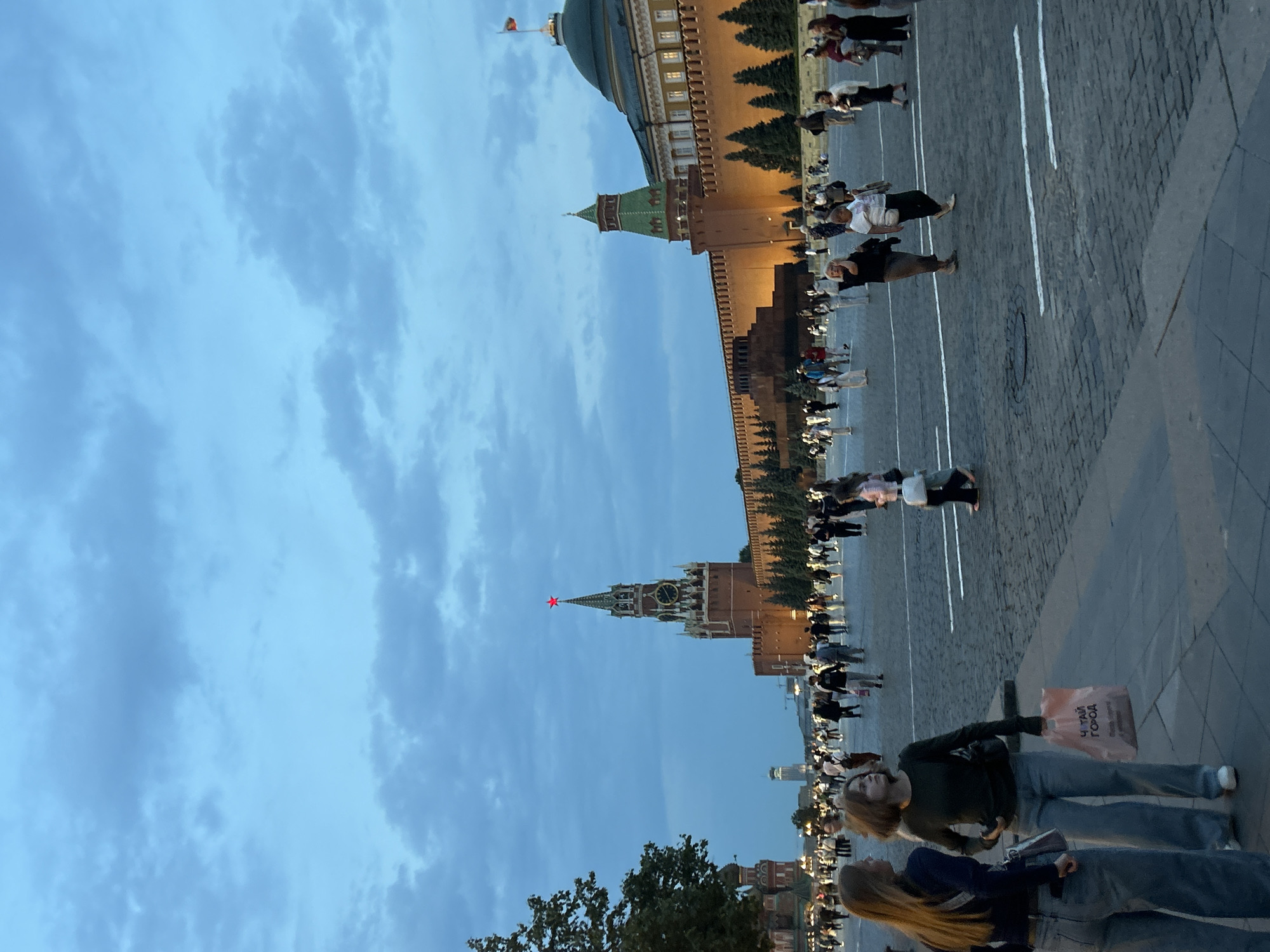 The image depicts a wide shot of Red Square in Moscow, Russia, during the twilight hours. The sky is a deep blue, with scattered clouds. Dominating the background is the red brick wall of the Kremlin, illuminated by artificial lights, and the iconic Spasskaya Tower with its red star topping the spire. To the right, a portion of the State Historical Museum is visible with its green dome. In the foreground, a paved plaza is filled with a crowd of people walking and standing. The lighting suggests it is either early morning or late evening. Two young women are in the immediate foreground on the left, facing away from the camera. One carries a pink shopping bag with Cyrillic text.