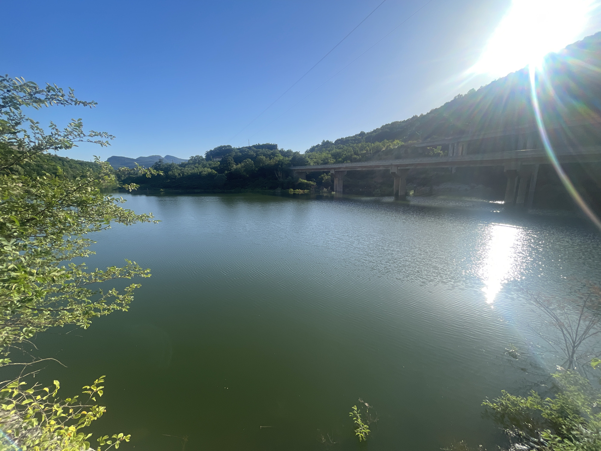 The image depicts a serene, sun-drenched waterscape with a clear blue sky. A calm, greenish lake fills the foreground and midground, with gentle ripples on its surface catching the sunlight. The sun is high in the sky, casting bright rays and lens flares, suggesting it is midday.
On the right side of the image, a sunlit hill covered in lush green trees slopes down towards the water. A concrete bridge, supported by large pillars, spans across the water in the midground, carrying what appears to be a road. The bridge and the surrounding hills are bathed in sunlight, highlighting the bright, warm weather.
On the left side, a cluster of green foliage from a tree branch extends into the frame, its leaves sharply defined against the bright sky. The overall scene evokes a sense of tranquility and natural beauty, with the man-made bridge integrated into the landscape. The location is identified as Asparuhovo, Bulgaria. No people or text are visible in the image.