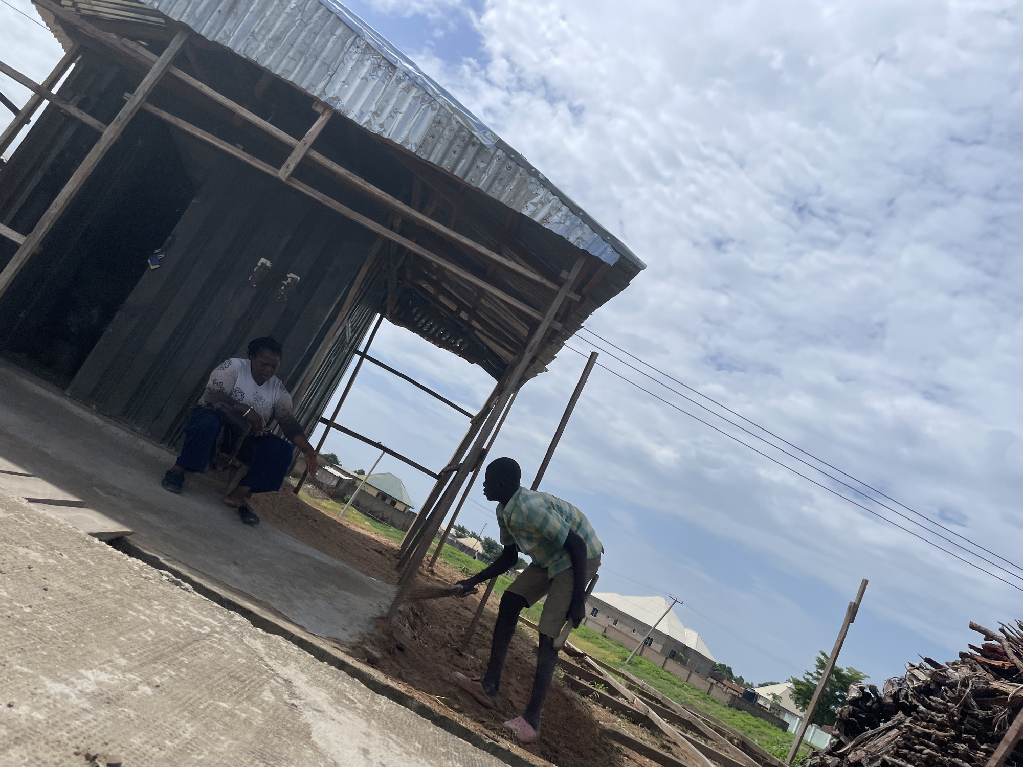 A construction scene in Jalingo, Nigeria, features two individuals working on a building. In the foreground, a young boy wearing a checkered shirt and shorts is sweeping the ground with a broom. To his left, a woman in a white patterned top and blue pants is crouched down, appearing to be observing or perhaps resting. The building under construction has a corrugated metal roof and a wooden frame. In the background, several houses and a pile of lumber are visible, suggesting a residential or developing area. The sky is cloudy, indicating it might be overcast or nearing a change in weather. The lighting suggests it is daytime. The overall mood appears to be one of labor and everyday activity.