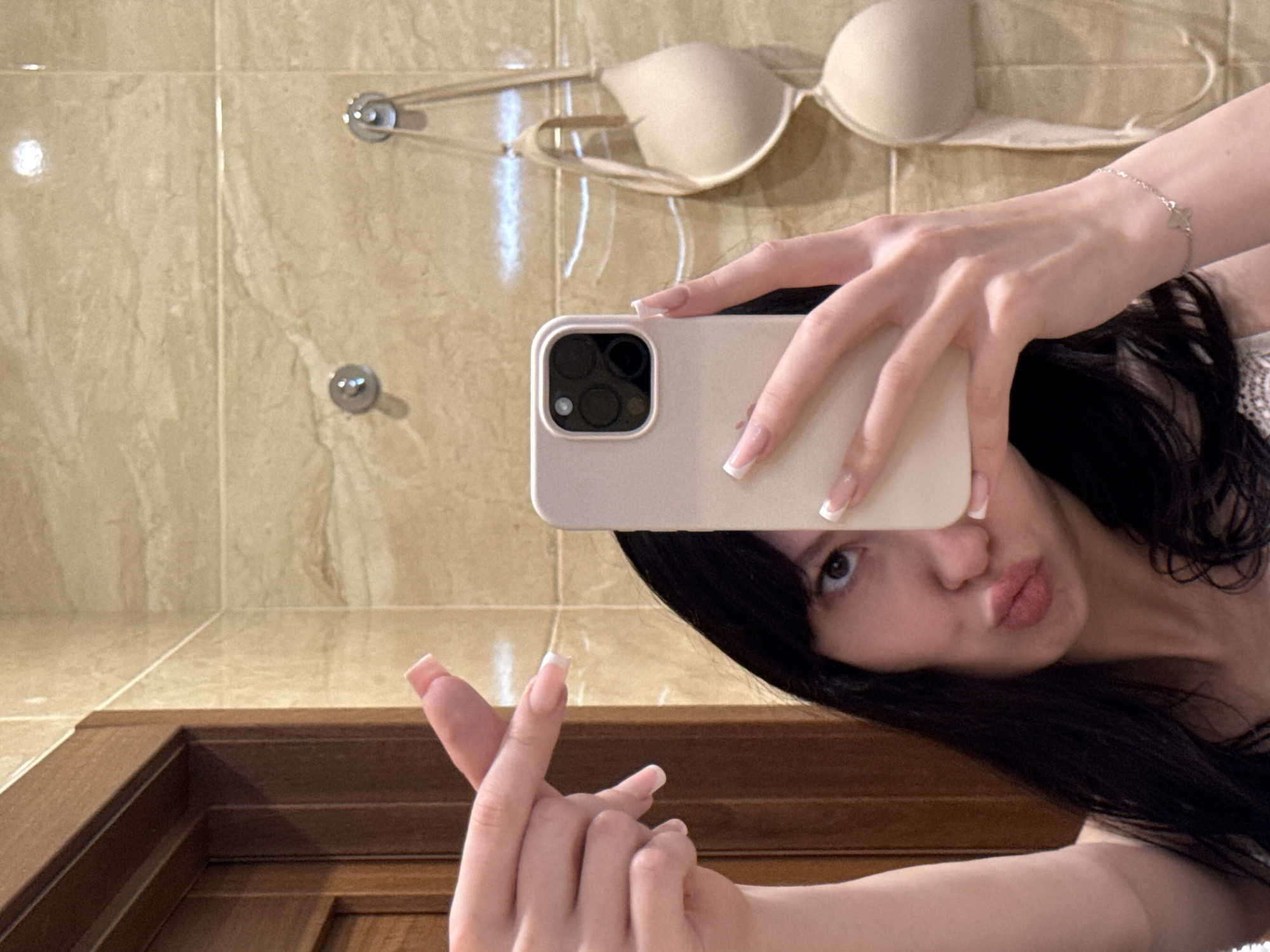 In a bathroom with beige marble tile walls and a wooden door, a woman with dark hair is taking a mirror selfie. She is wearing a white lace top and has a light-colored phone in her right hand. Her left hand is raised with her index and middle fingers extended. Two bras are hanging on hooks on the wall behind her. The scene is located in Khimki, Russia.