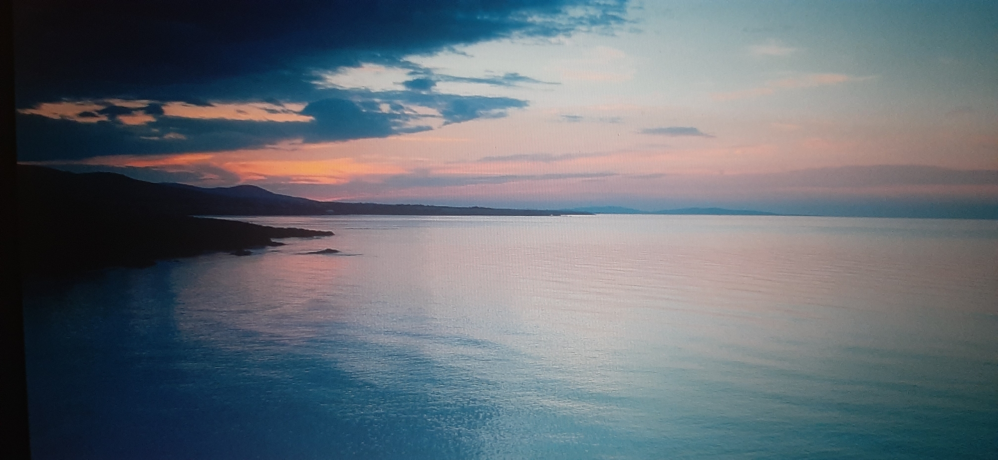 The image depicts a serene coastal landscape at sunset or sunrise. The sky is a canvas of soft hues, transitioning from deep blues and grays at the top to warm oranges and pinks closer to the horizon. Wispy clouds are scattered across the sky, catching the fading light. In the foreground, a calm body of water reflects the colors of the sky, with gentle ripples disturbing its surface. To the left, a dark, silhouetted coastline with rolling hills stretches into the distance. The overall impression is one of tranquility and natural beauty. There are no people, animals, or man-made structures visible in the scene. The location is not Yessentukskaya, Russia; this image is of a coastal area.