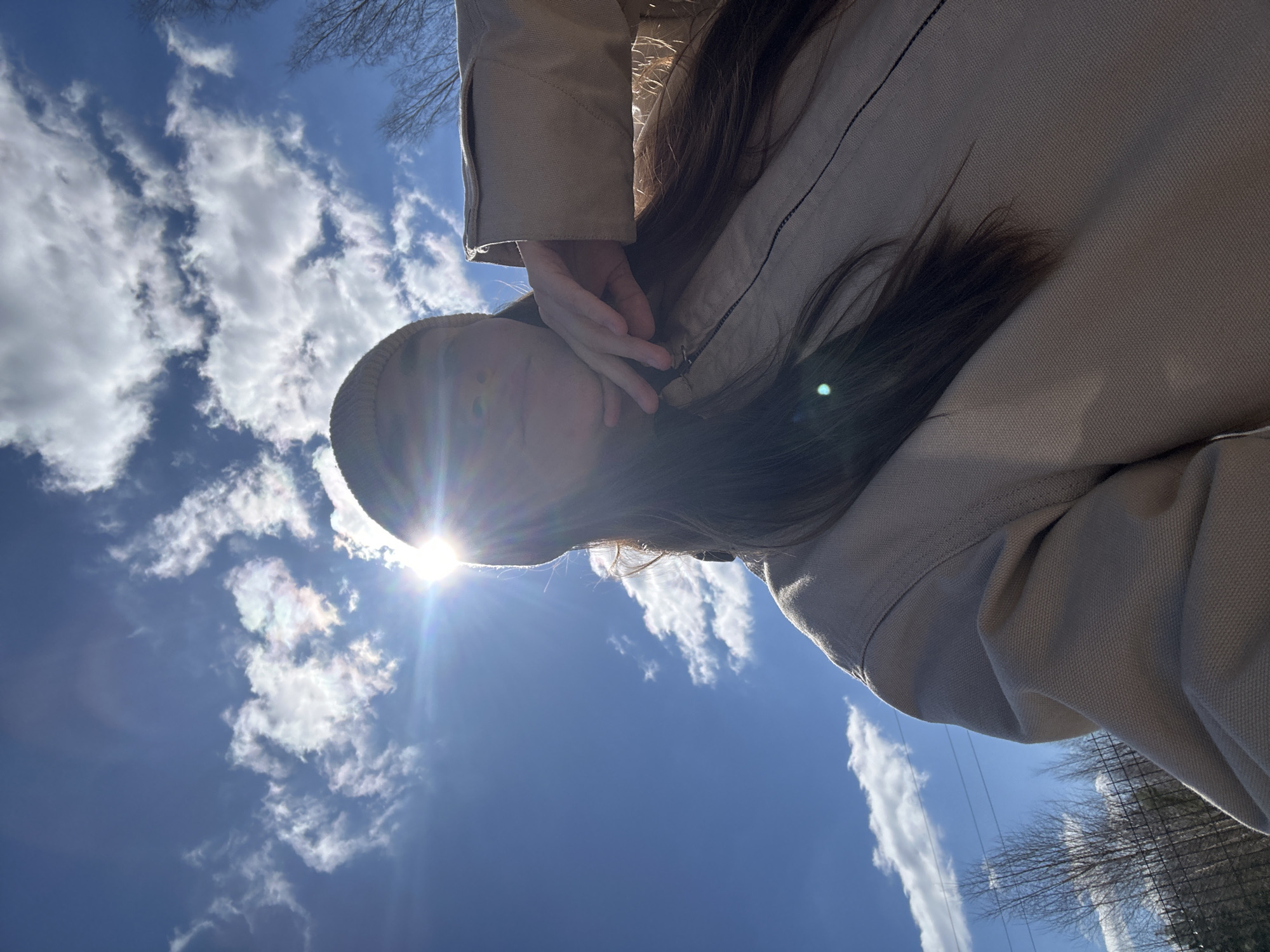 A low-angle shot captures a person with long, dark hair wearing a tan jacket and beanie. Their hand is raised near their face, as if shielding their eyes. The sun shines brightly behind their head, creating lens flare. The background features a blue sky filled with puffy white clouds. Bare tree branches are visible in the corners of the frame. No information is available to determine the location as the city of Uva, Russia.