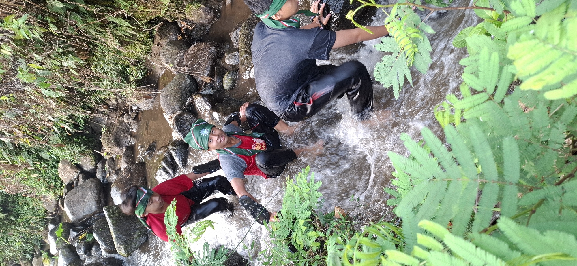 In the natural environment of Puntukrejo, Indonesia, three young boys are wading through a shallow, rocky stream. The water flows over dark, smooth stones, and lush green foliage lines the banks and encroaches into the stream itself. The boys are dressed in athletic wear, with two wearing red shirts and one a dark gray shirt. All are also wearing dark, possibly black, pants that are wet and clinging to their legs. They are also wearing green headbands with a white pattern, and one boy has a red scarf around his neck. The boy in the center of the image is looking at the camera and smiling, while the other two appear focused on navigating the stream. The boy on the left is holding a black shoe in his hand, and the boy on the right is reaching for a rock. The overall scene suggests an outdoor activity or adventure, possibly a hike or a camping excursion.