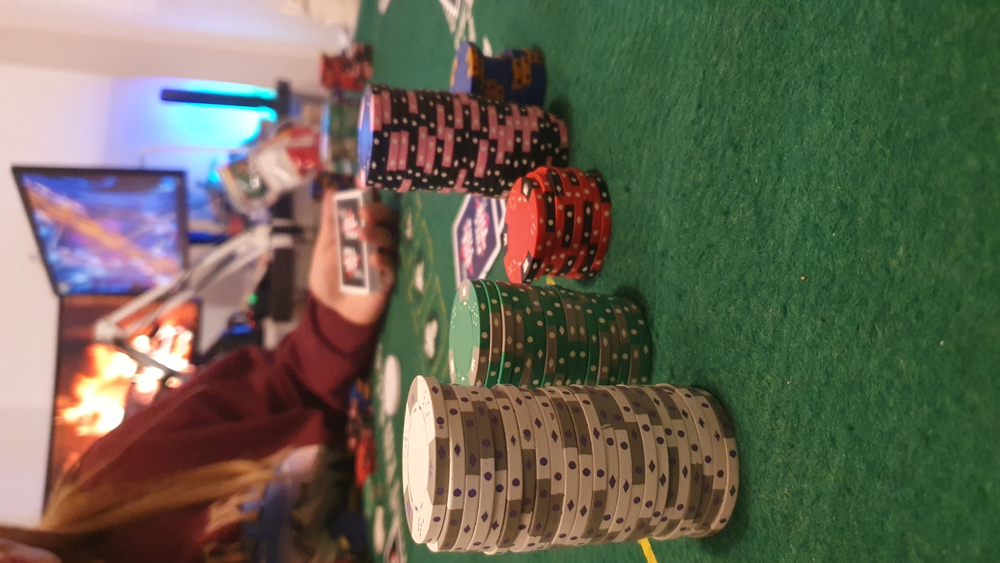 The image shows a poker game in progress. There are stacks of poker chips in various colors including white, green, red, black and pink, and blue. A person is holding up playing cards. A poker mat is present. In the background, there are computer monitors with images displayed and colored lighting.