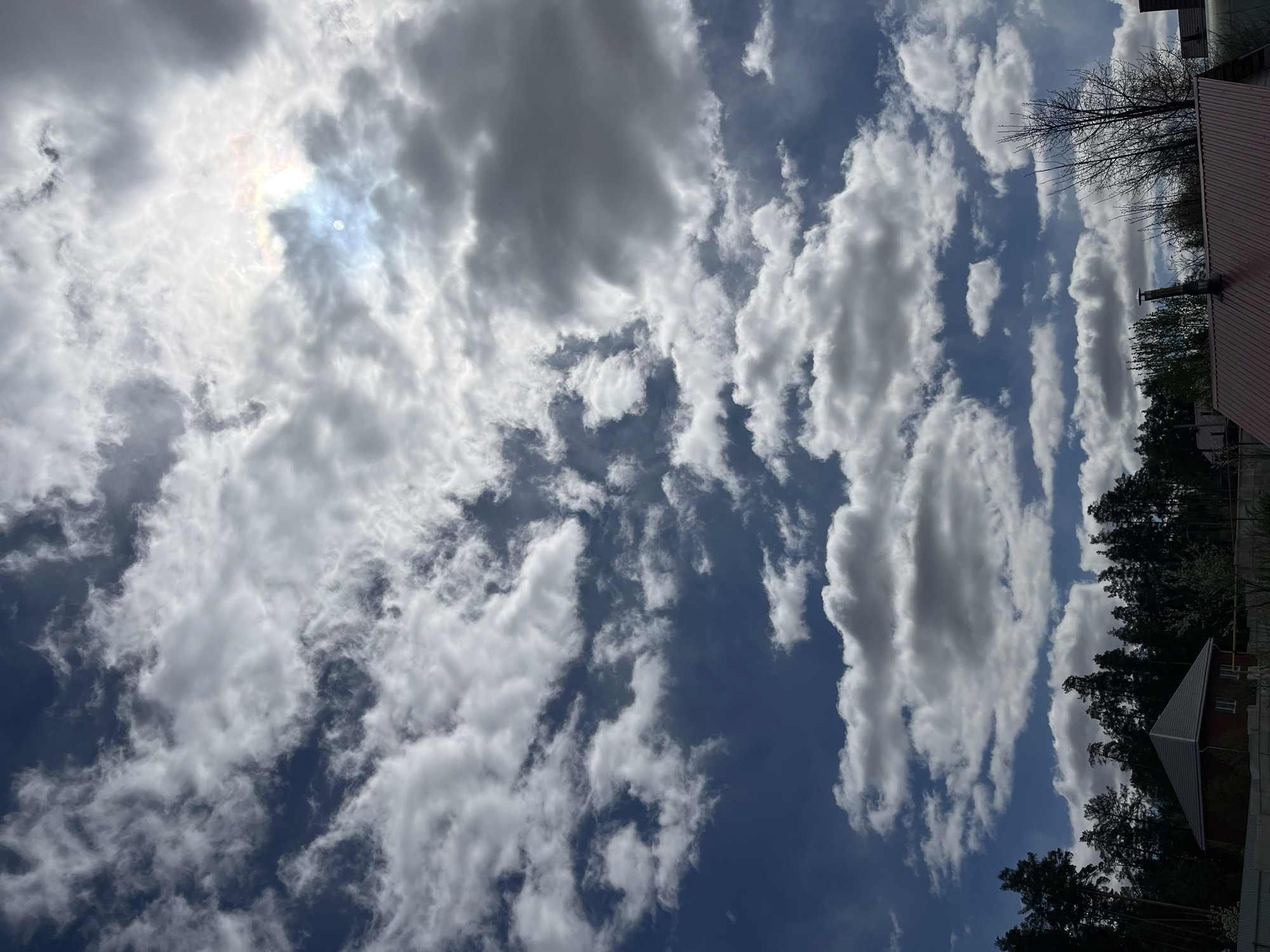 The image shows a view looking up at a sky filled with white and gray clouds, set against a backdrop of blue sky. The sun is visible behind some of the clouds, producing a bright glow. Part of a building and trees are visible on the right side of the frame. The image was taken in the city of Chaikovsky, Russia.