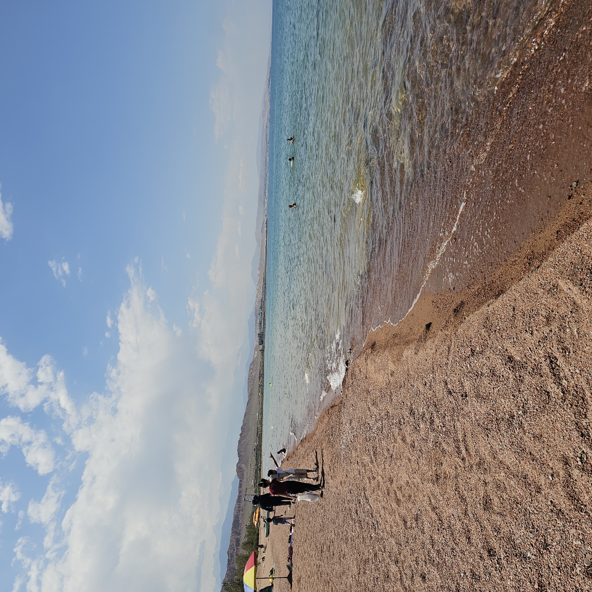 This is a ground-level, wide-angle photograph of a beach on the shore of Issyk-Kul Lake, near Kaji-Say, Kyrgyzstan. The beach is composed of small, reddish-brown pebbles and sand. In the foreground, to the left, a colorful umbrella shades a tent and some items laid out on the beach. A group of people are walking along the shoreline, some carrying bags. Further down the beach, a few individuals are visible near the water. In the middle ground, several people are swimming and wading in the clear, turquoise water. The background features a distant shoreline with buildings and a range of rugged, arid mountains under a bright blue sky with scattered white clouds. The water is calm with gentle waves lapping the shore.