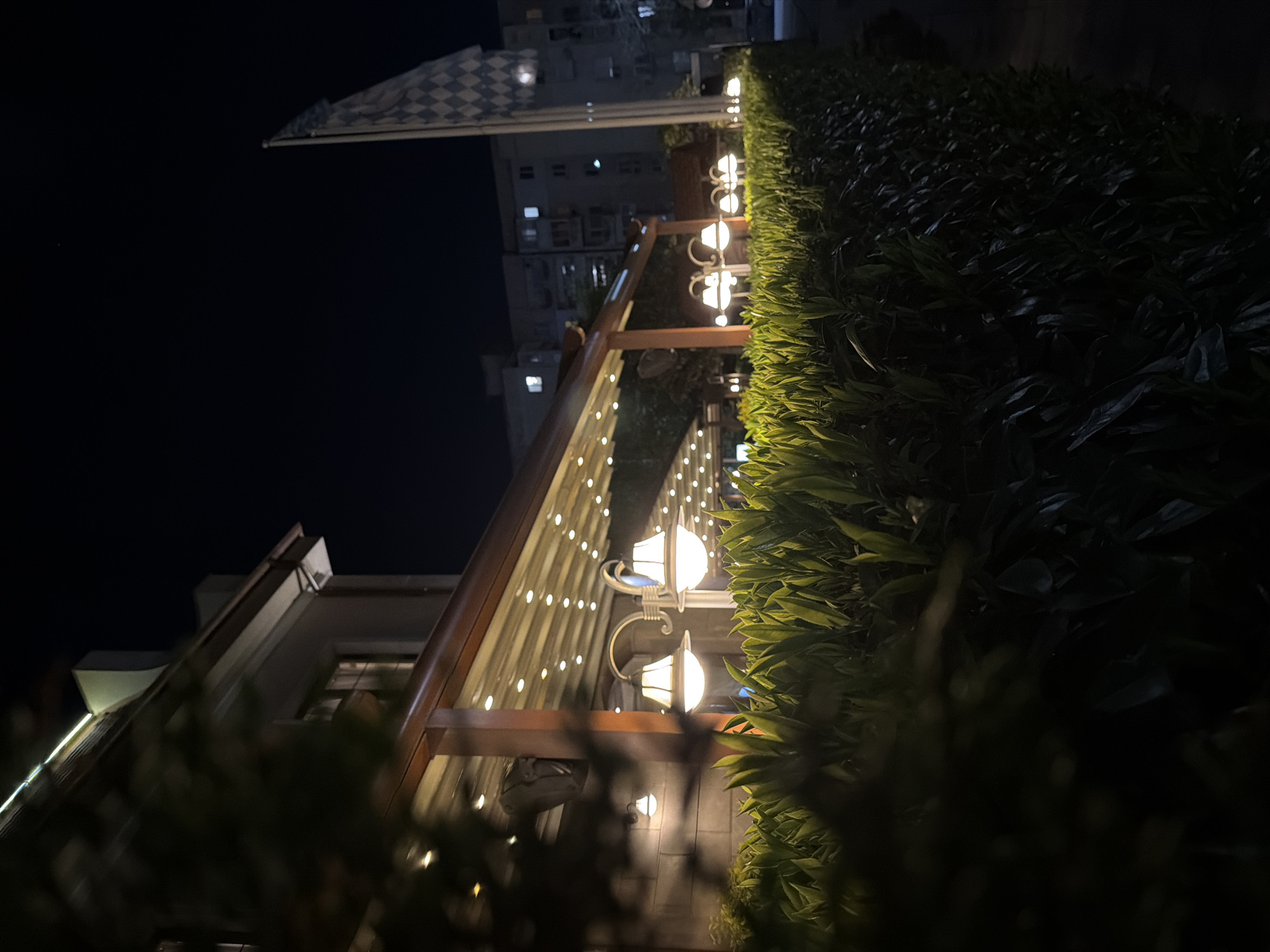 The image presents a vibrant nighttime outdoor scene, likely an illuminated patio or garden area of a building, possibly a restaurant or cafe. The dark night sky dominates the upper portion of the frame, providing a stark contrast to the brightly lit elements below.
A prominent feature is a covered outdoor structure, possibly a pergola or awning, adorned with numerous warm-toned string lights that create a festive glow. Along the right side of the frame, a dense, manicured hedge or row of bushes is richly illuminated by several ornate, traditional-style wall-mounted lamps. These lamps are spaced along a wall or fence, casting a soft light on the lush green foliage.
In the background, a multi-story building with some illuminated windows is visible against the dark sky, suggesting an urban or suburban setting. A distinctive checkered flag, featuring a white and dark grey/black pattern, flies from a pole in the upper left corner of the image, adding an intriguing detail.
The overall atmosphere conveyed is one of warmth, coziness, and invitation, characteristic of an evening spent in a pleasant outdoor setting. No people or specific activities are discernible, and no text is visible within the frame. The image emphasizes artificial lighting and natural greenery against the night. The time of day is clearly night, and the weather appears clear.