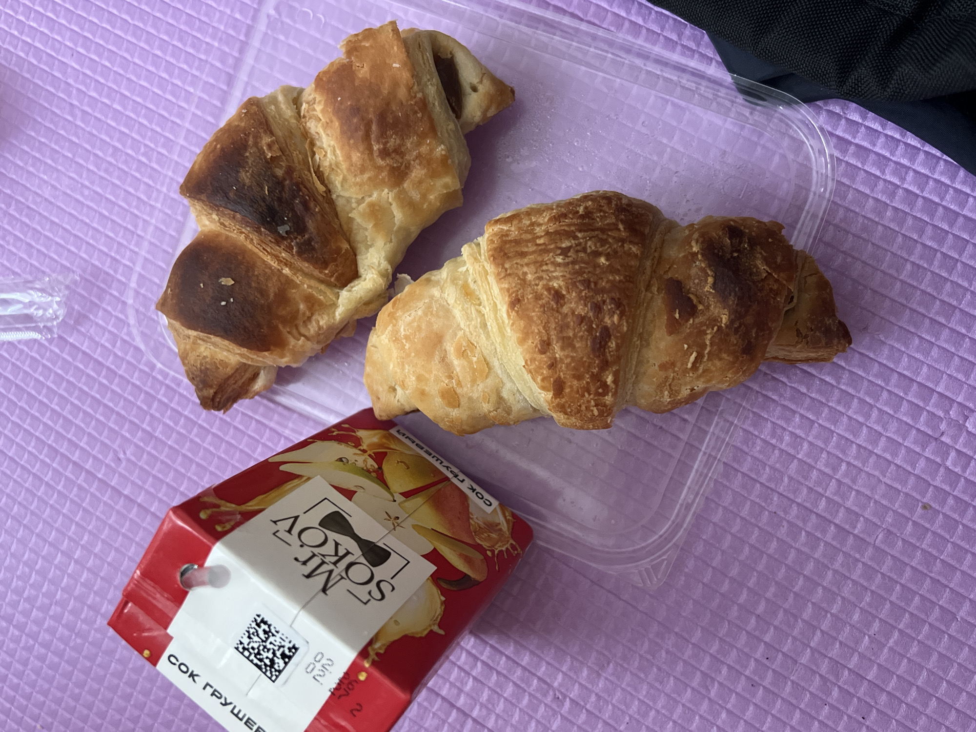 The image shows two croissants in a clear plastic container, next to a red and white box of Mr. SOKOV pear juice. The setting appears to be indoors, with a purple, textured surface beneath the items. There is no clear indication of the location being in Berezniki, Russia within the image.