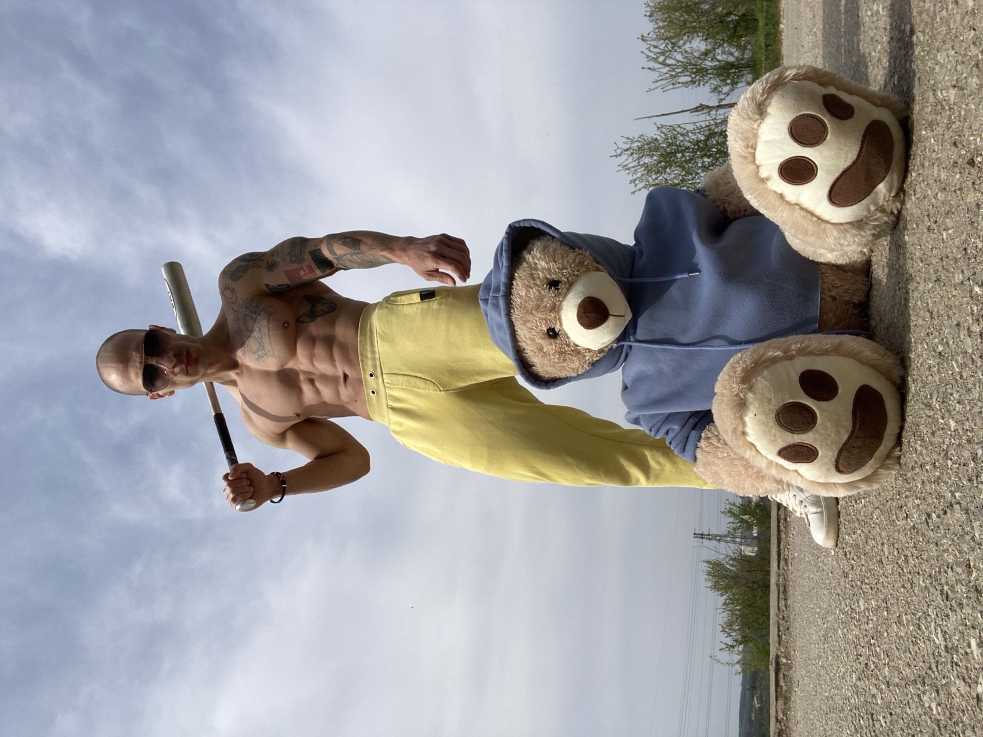 A shirtless, muscular, bald man with tattoos, wearing sunglasses and yellow pants, is holding a baseball bat. He stands next to a large teddy bear wearing a blue hoodie and white sneakers. The setting appears to be an outdoor concrete area near trees and power lines, located within the vicinity of Pleven, Bulgaria.