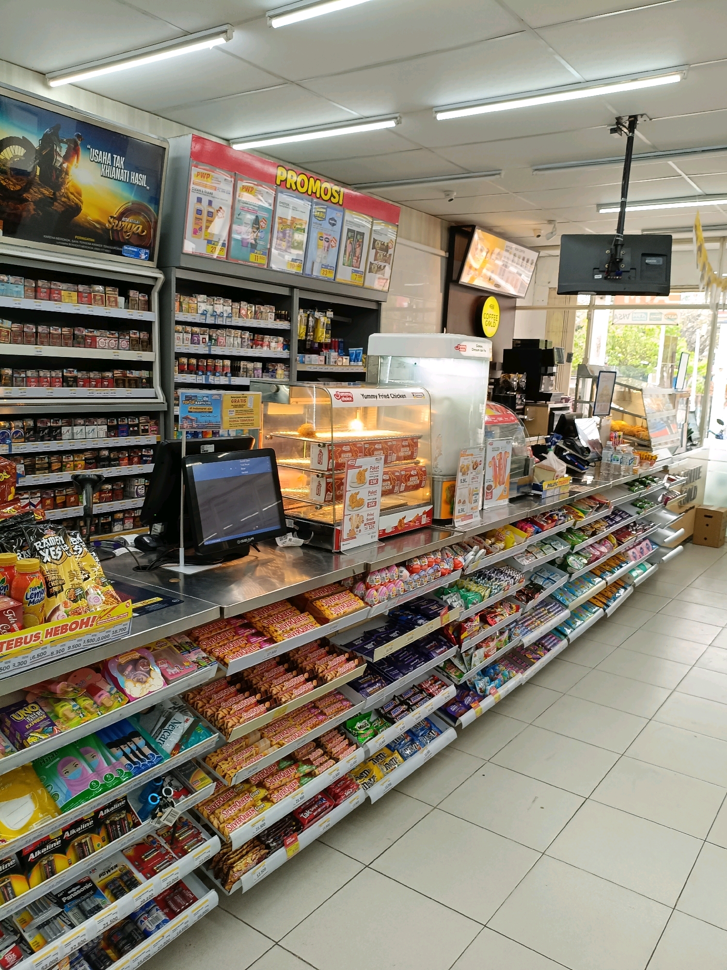 The image depicts the interior of a convenience store in Maguwoharjo, Indonesia, specifically a section of the store featuring a checkout counter and adjacent shelves stocked with various consumer goods. A computer monitor, point-of-sale system, and a hand-held scanner are present on the metallic countertop of the checkout area. Behind the counter, a heated display case contains fried chicken and other food items.
The shelves on the right side of the image are filled with an assortment of confectionery, including chocolate bars, candies, and snack items. Some shelves also display batteries and other packaged goods. The store's ceiling is illuminated by fluorescent lighting. Promotional posters are visible on the walls and above the shelves, advertising various products and discounts. The floor is tiled with light-colored square tiles. The overall impression is of a well-stocked and organized retail environment.
