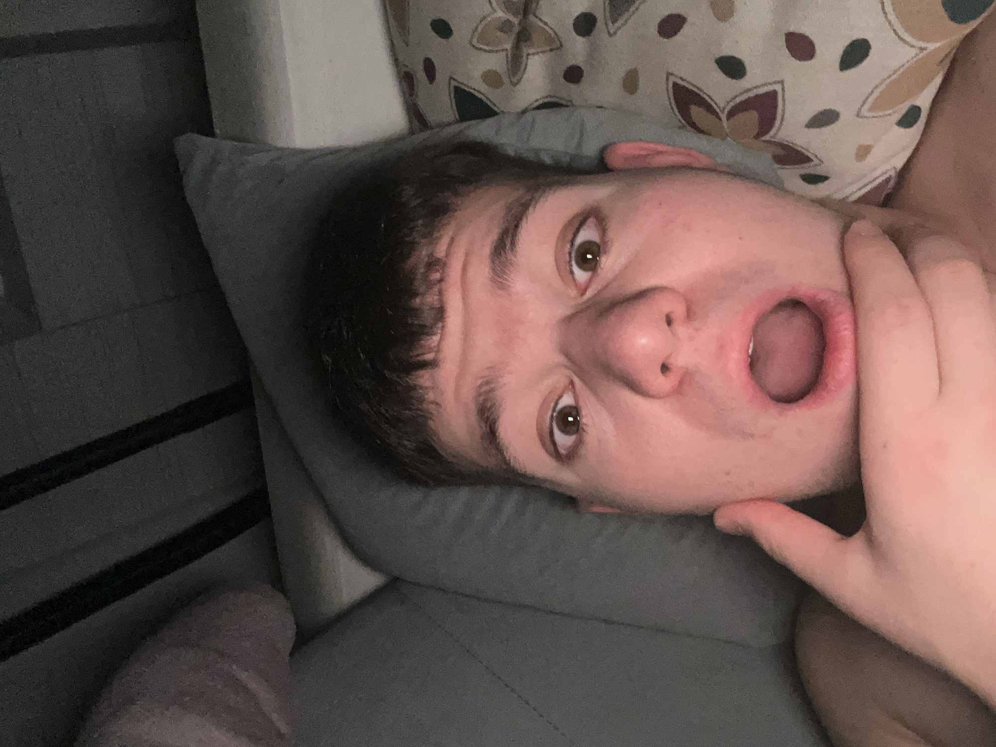A close-up, poorly lit photograph shows a young man lying on a gray pillow with a patterned pillow behind him. He has dark hair and brown eyes, and his mouth is open. His hand is touching his face. The environment includes bedclothes and slats of a bed frame. The location is not discernible from the image.