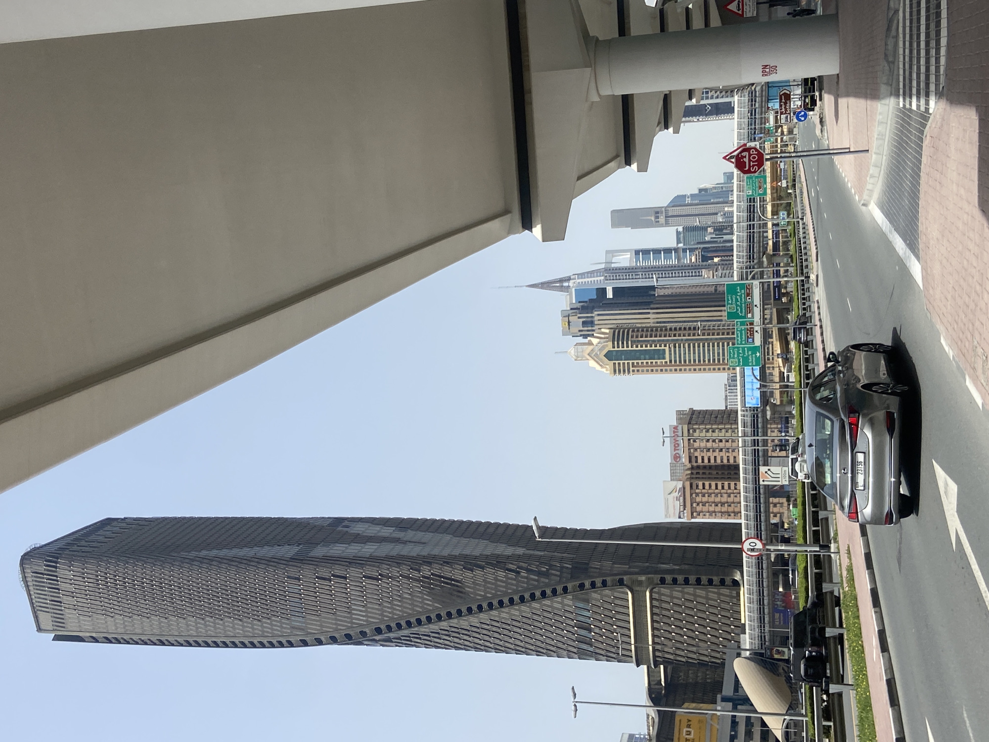 The image shows a street scene in Dubai, United Arab Emirates. In the foreground, the architecture of a modern skyscraper dominates the view, featuring a distinctive geometric pattern on its facade. A bridge structure is visible at the top left corner of the frame, providing a partial covering over the road. The road has at least three lanes and a grey car is present on the right side, parked along the roadside. Street signs and traffic signals line the road. In the background, a cluster of other skyscrapers forms the city skyline. The sky is light blue.