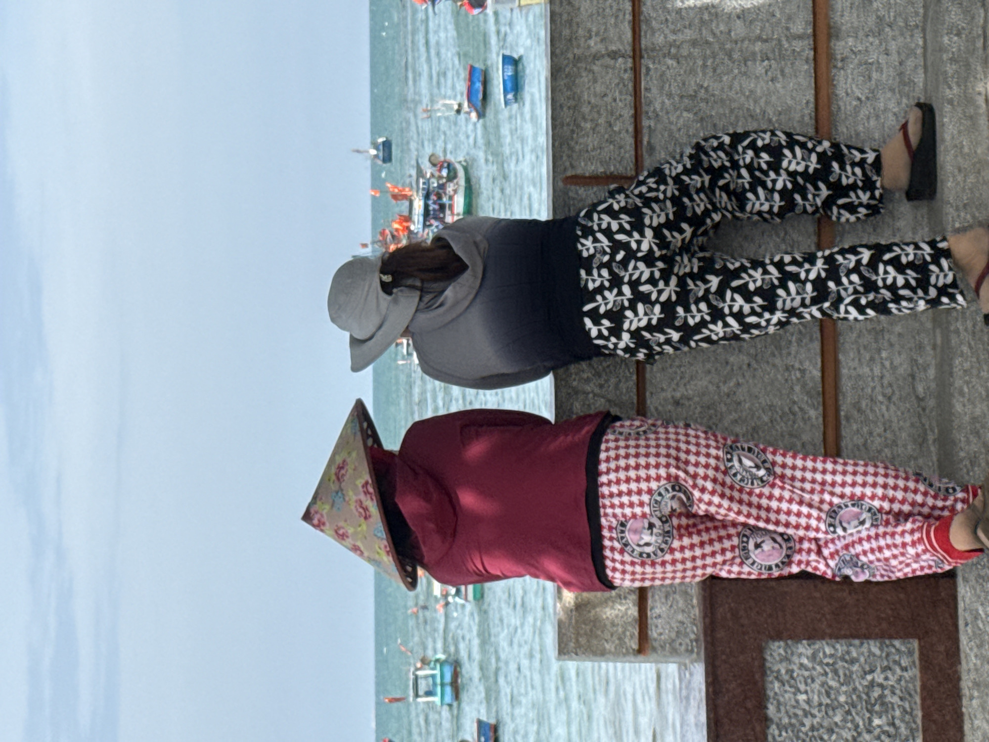 The image depicts two individuals from behind, standing on a stone structure overlooking a body of water, likely the sea, with numerous small boats scattered across the surface. The sky is overcast with pale blue clouds. The person on the left wears a conical straw hat with a floral pattern, a dark red hooded jacket, and loose-fitting red and white checkered pants with circular emblems. The person on the right wears a wide-brimmed grey hat, a grey hooded jacket that fades to black at the bottom, and loose-fitting black pants with a white leaf pattern. They are both looking out at the water. The location is specified as Vĩnh Lương, Vietnam.