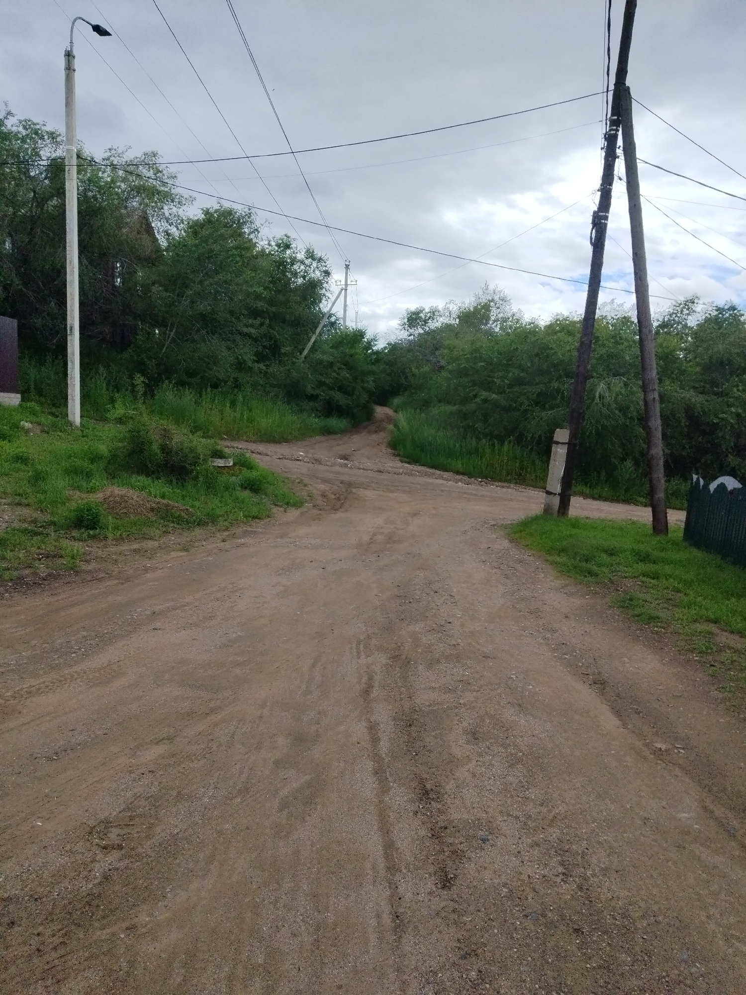 The image depicts a dirt road in Chita, Russia, likely in a rural or semi-rural area, given the presence of vegetation and utility poles. The road is unpaved, with visible tire tracks and loose gravel. It curves into the distance, surrounded by dense green foliage and trees on both sides.
Dominating the left side of the frame is a concrete utility pole with a street light fixture at its top, adorned with a black lamp. Further in the background, another utility pole can be seen. On the right side, a pair of weathered wooden utility poles stand closely together, supporting numerous electrical wires that crisscross the sky. Near these poles, a section of a dark green fence is visible.
The sky is overcast with a thick layer of clouds, suggesting a dull or potentially rainy day. The overall impression is one of a simple, somewhat neglected, outdoor setting.