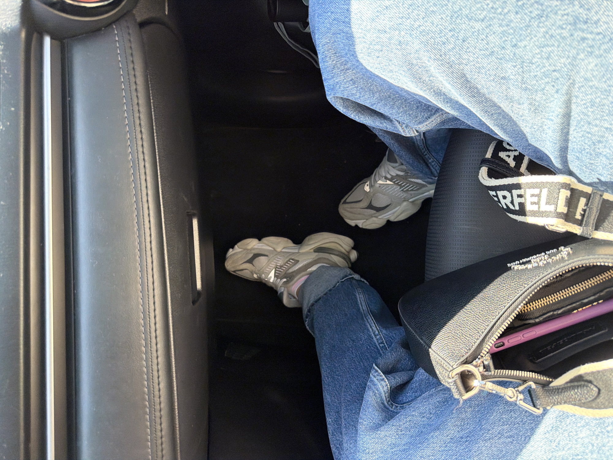 A person is seated in a vehicle. They are wearing blue jeans and gray sneakers. A black handbag with a Karl Lagerfeld strap rests in their lap, partially open, revealing a purple cell phone. The interior of the vehicle is primarily black, featuring a leather-covered seat. The location is impossible to determine based on the provided image.