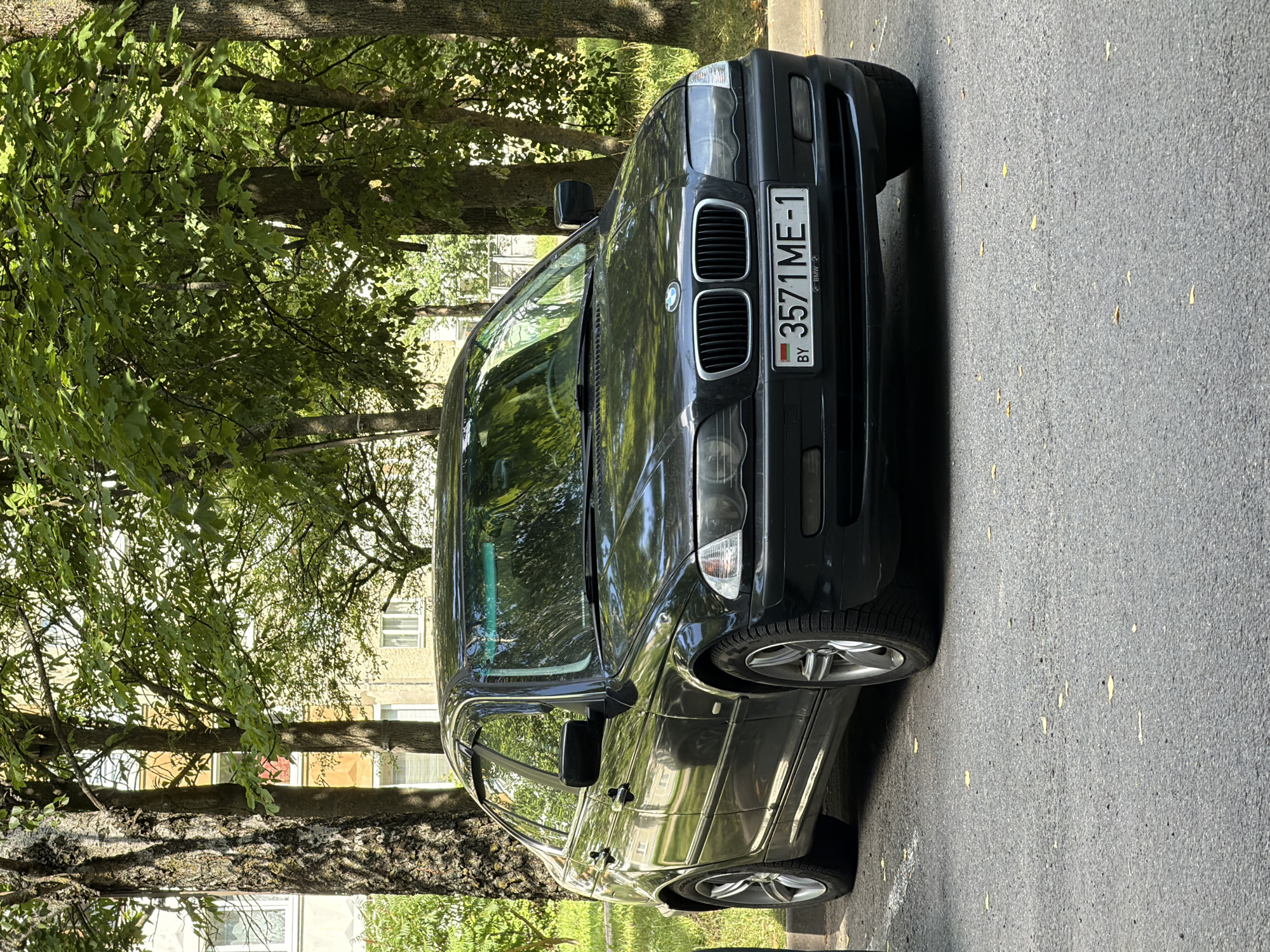 A black BMW sedan is parked on an asphalt surface in front of a residential building with a pale yellow facade and cream-colored trim. Trees with dense green foliage are situated between the building and the car, casting shadows on the vehicle and the surrounding area. The car's license plate reads "3571ME-1" with "BY" indicating Belarus. The scene is set in Minsk, Belarus, under daylight conditions. No people are visible in the image.