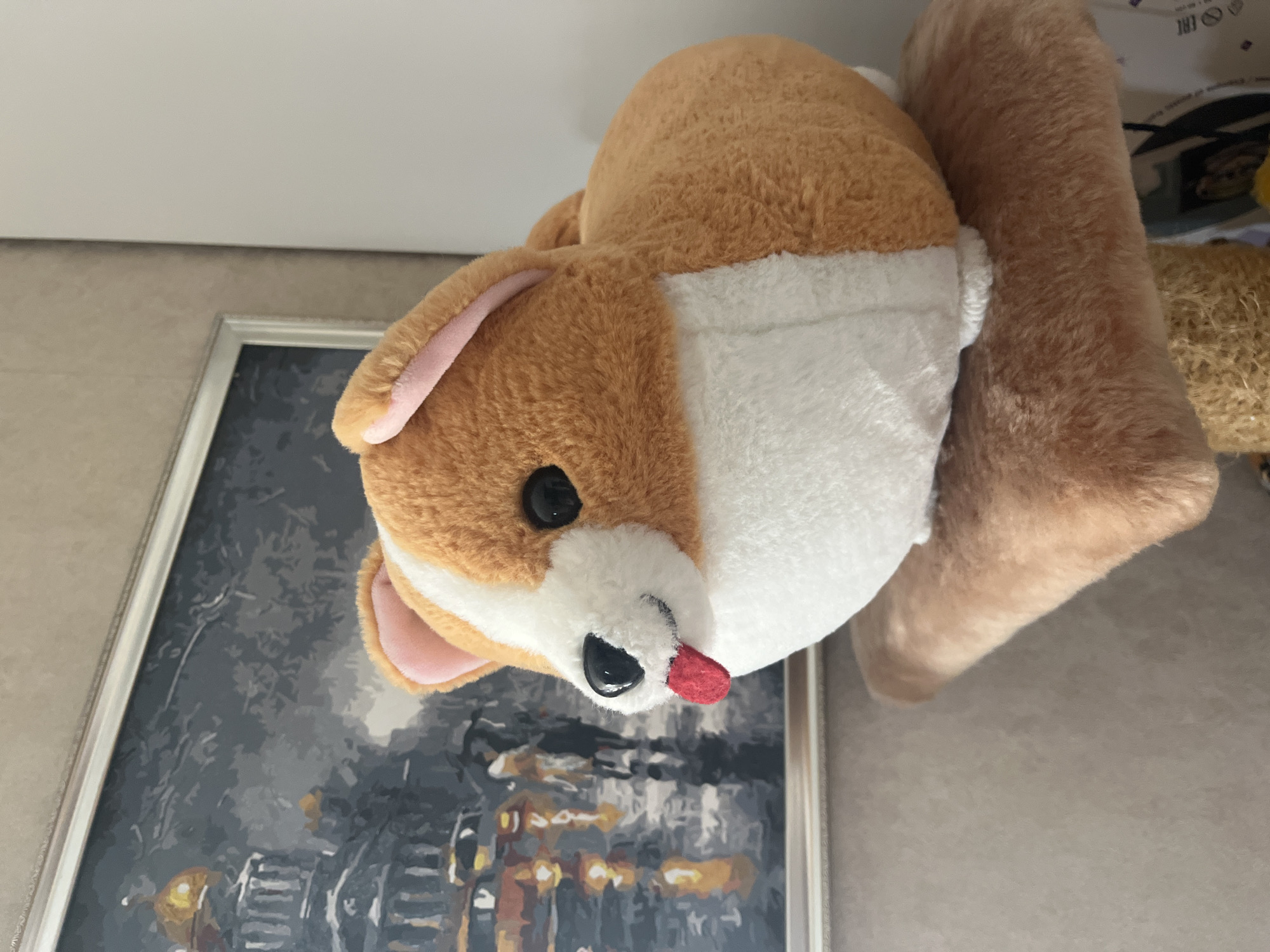 A close-up, medium shot captures a plush Corgi toy with its tongue sticking out. The toy is light brown and white, with black eyes and pink inner ears. It sits on a fuzzy brown platform, which appears to be part of a cat tree or similar furniture. Behind the toy, a framed painting depicts a rainy cityscape with blurred figures holding umbrellas and reflections of lights on a wet street. The painting has a metallic frame. The background wall is textured and cream-colored on the right. The scene does not contain any recognizable landmarks or elements that could identify its location in Vladimirovka, Russia.