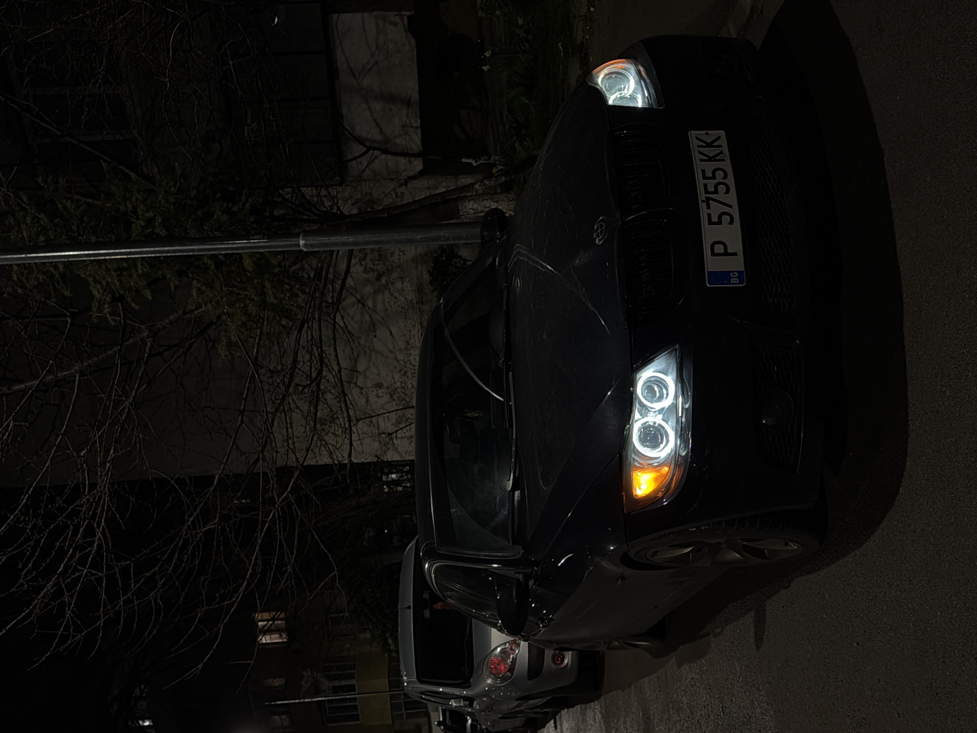 A nighttime street scene in Varna, Bulgaria, features a dark-colored BMW 3-series (E90) sedan parked on the right side of an asphalt road. The BMW's front is angled towards the viewer, displaying illuminated white "angel eye" headlights and an amber turn signal active on its passenger side (viewer's left). The front license plate, "BG P 5755 KK," confirms registration in Varna. A silver Renault Kangoo van is parked immediately behind the BMW. The background on the left reveals a multi-story building with several illuminated windows. To the right, bare-branched trees and a vertical street lamp pole are visible against a darker building or wall. The road surface appears dark and possibly damp. No individuals are present in the image.