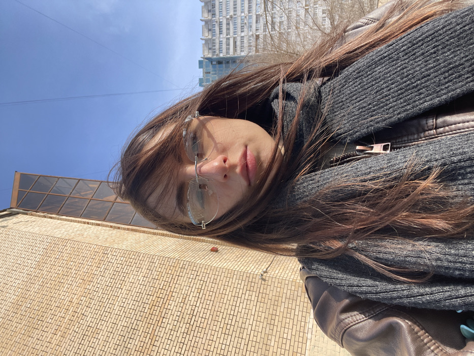 A female individual is depicted in a medium-shot selfie, positioned outdoors in an urban environment in Moscow, Russia. Her long brown hair, styled with bangs, is partially wind-swept across her face. She wears rimless glasses with light-tinted lenses, a brown leather jacket, and a dark grey ribbed knit scarf. Her gaze is directed towards the camera. The background on the left features a light-colored brick building with a vertical, brown-tinted glass structure extending upwards. A small red fixture is present on the brick wall. The background on the right reveals a clear blue sky and distant multi-story residential buildings, some with light facades and others with darker, possibly reflective, windows. Sparse, bare trees are visible between the subject and the distant buildings. The scene is illuminated by bright sunlight.