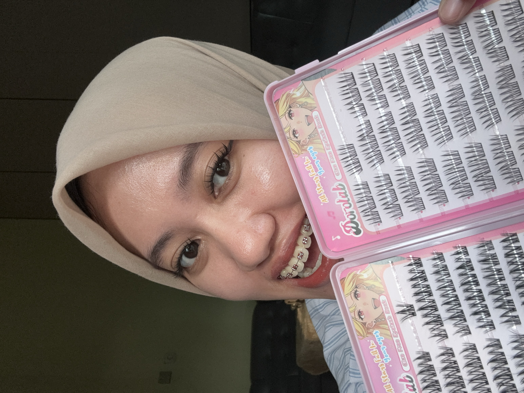 The image shows a woman wearing a light beige hijab, smiling and displaying two pink plastic cases of "Dorolab All Stars Fall In Your Eyes" glue-free individual false eyelashes. She has braces on her teeth. The photo appears to be taken indoors, with a dark green wall visible in the background. While the location in the city of Gadingrejo, Indonesia is mentioned, it is not discernable within the image itself.